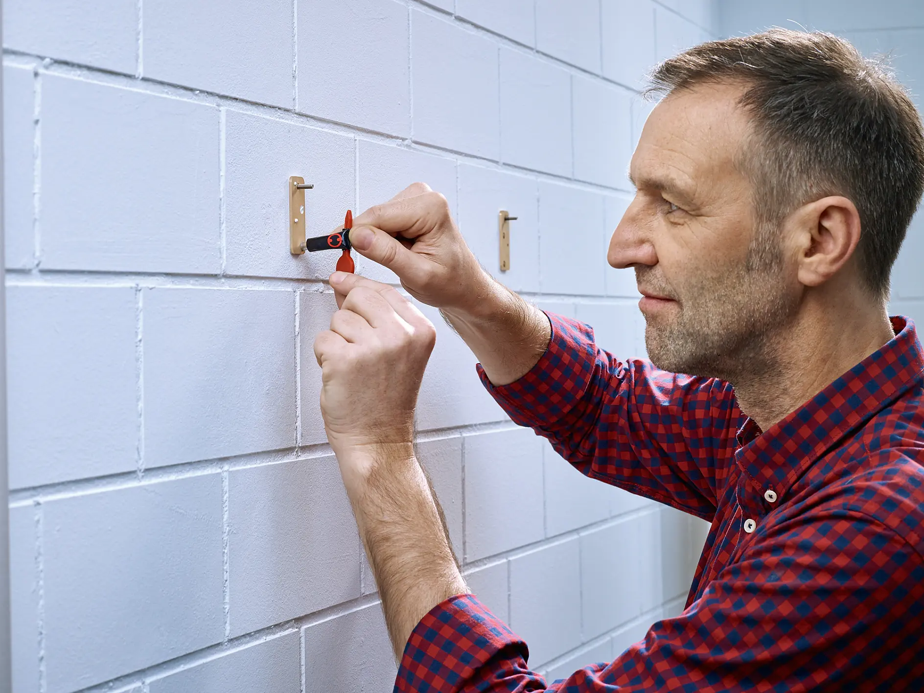 A man in a red and blue checkered shirt uses a flathead screwdriver to secure a bracket to a white brick wall. He is focused on tightening the screw, and the wall shows two installed brackets with tesa tape. (This text has been generated by AI)