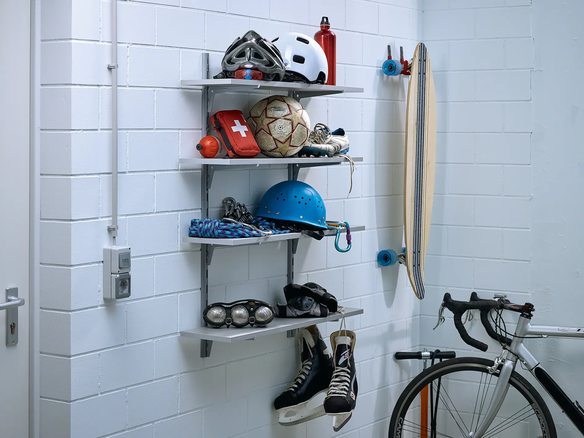 Wall-mounted shelves holding various sports equipment, including a climbing helmet, sports balls, a first aid kit, rollerblades, and a water bottle. A skateboard is hung on the wall using tesa tape, and a bicycle is parked nearby. (This text has been generated by AI)