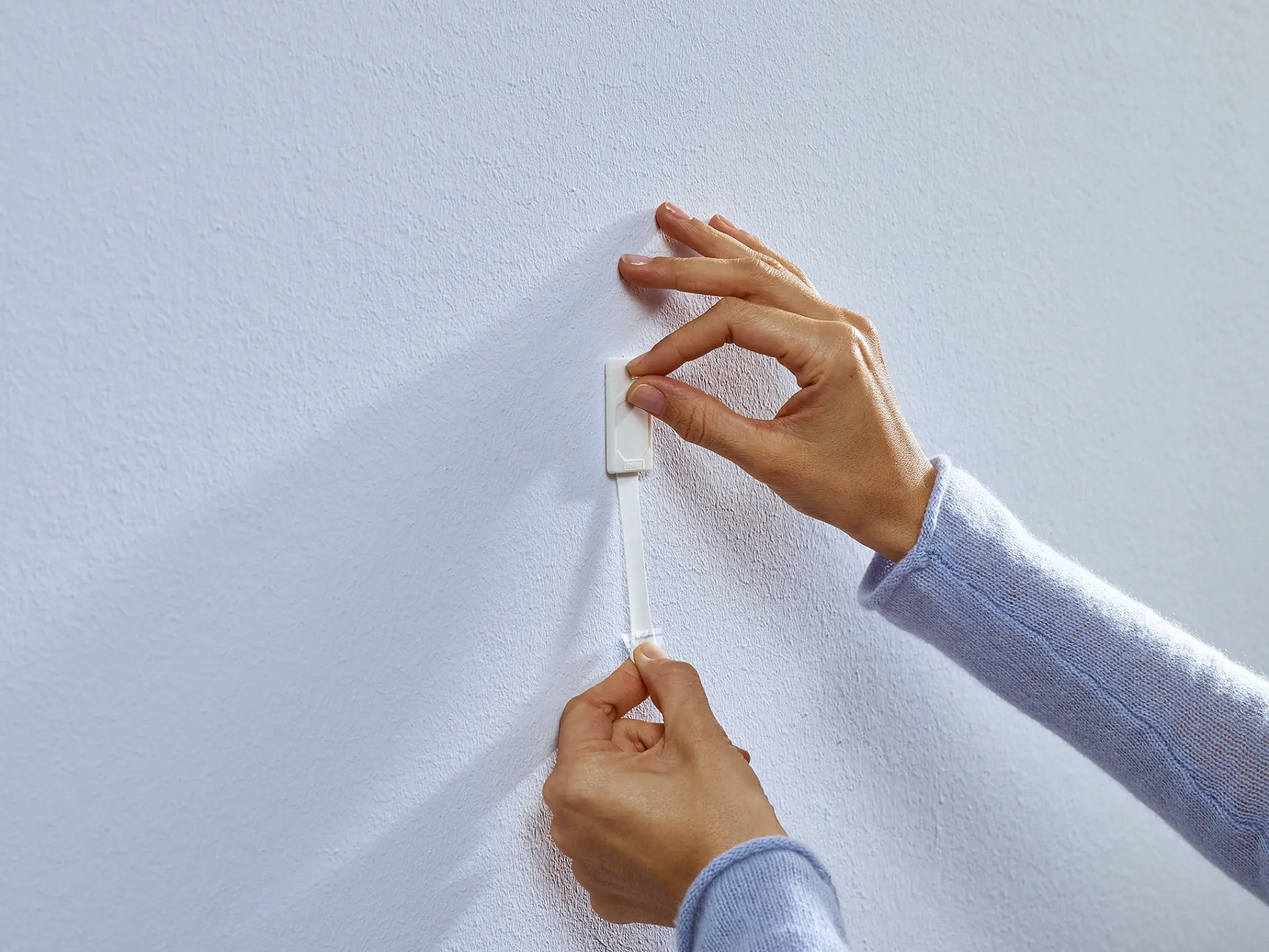 Two hands are attaching a white hook with tesa tape backing to a textured white wall. The person is pressing the hook firmly with one hand and securing the tesa tape strip with the other. The person is wearing a light blue long-sleeve top. (This text has been generated by AI)