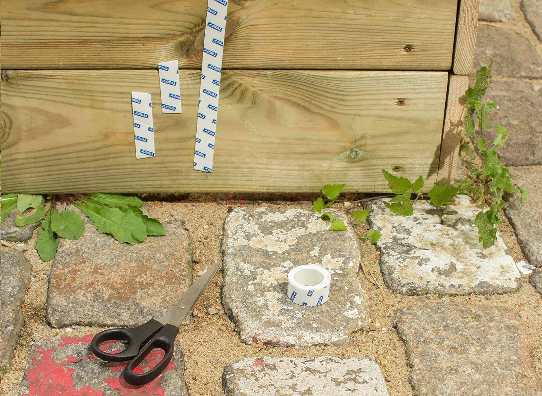 Scissors, tesa Powerbond® OUTDOOR adhesive tape and strips of tape applied to sandbox.