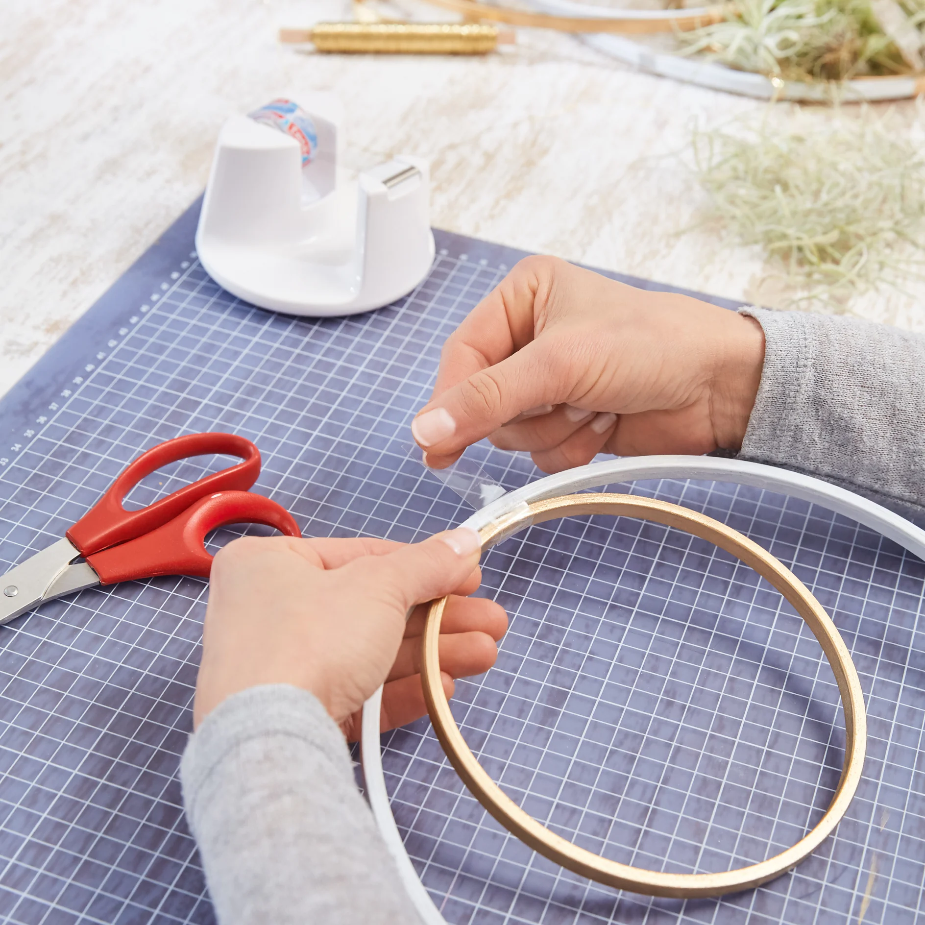 Hands are seen working on a craft project using a gold hoop and transparent tesa tape on a gridded blue mat. Red scissors and a tesa tape dispenser are nearby, with some greenery in the background. (This text has been generated by AI)