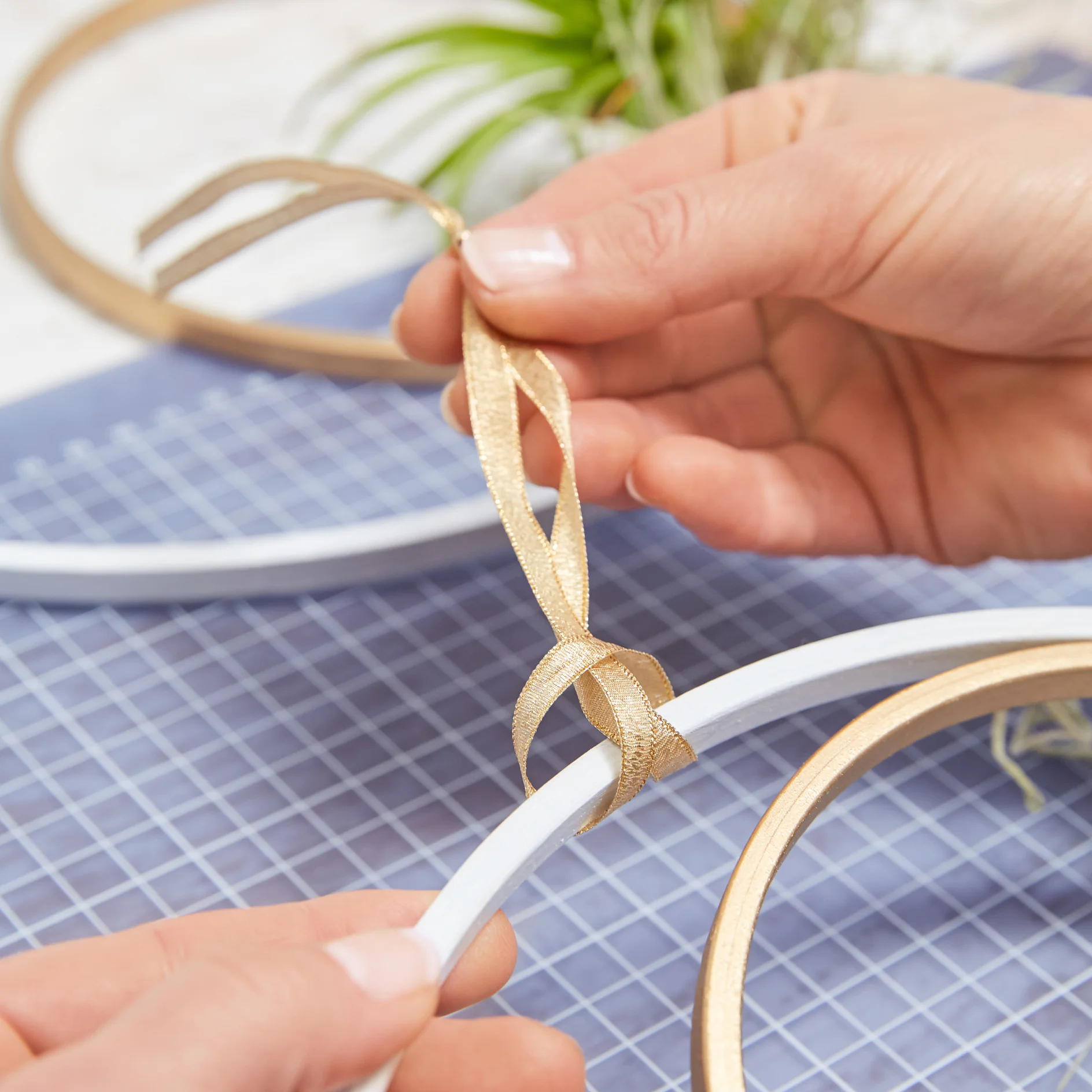 Two hands are tying a gold ribbon around a wooden hoop. The hoop is partially wrapped in white tesa tape. The scene is set on a blue grid mat with another hoop and some green foliage in the background. (This text has been generated by AI)