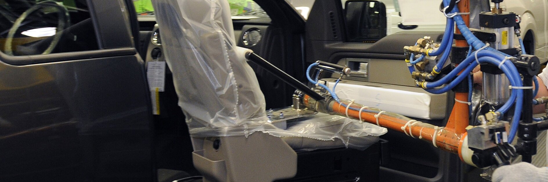 Une machine de montage installe un siège recouvert de plastique à lintérieur dun véhicule. La porte du véhicule est ouverte et divers bras mécaniques équipés de câbles et de tubes sont visibles, ce qui indique un processus dinstallation automatisé dans une usine de fabrication utilisant du ruban adhésif tesa. (This text has been generated by AI)
