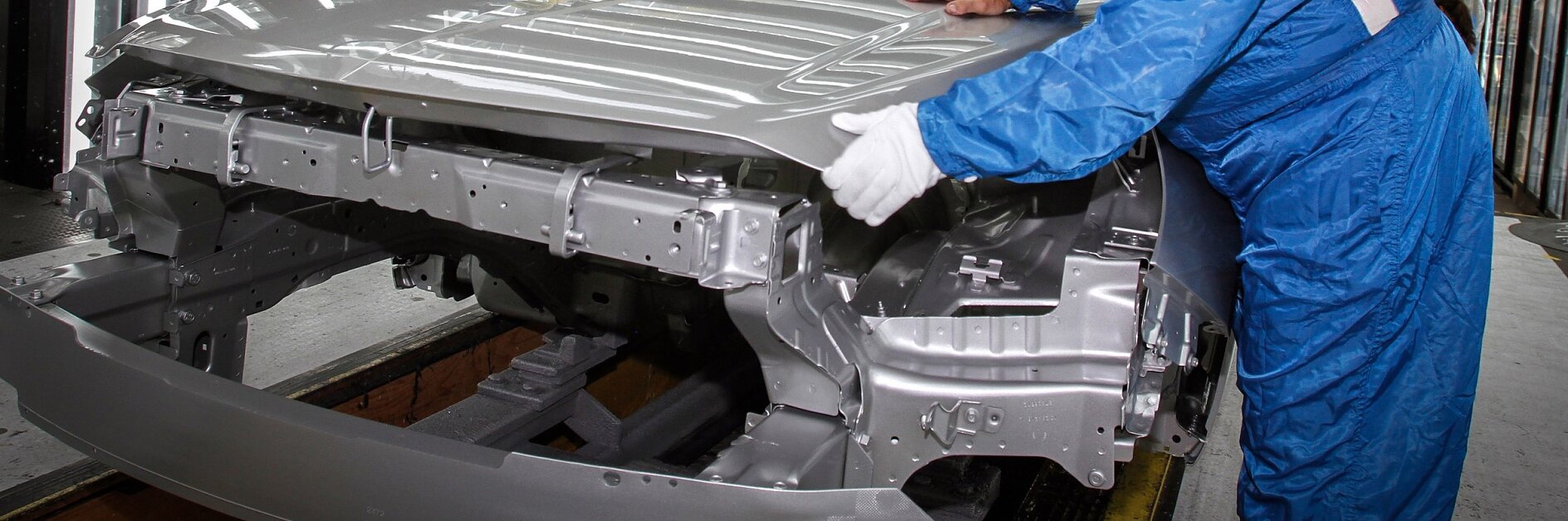 Une personne en combinaison bleue et gants blancs manipule un châssis de carrosserie argenté sur une chaîne de montage automobile. Le décor semble être celui dune usine ou dun site de fabrication, où le ruban adhésif tesa peut être utilisé pour diverses tâches dassemblage. (This text has been generated by AI)