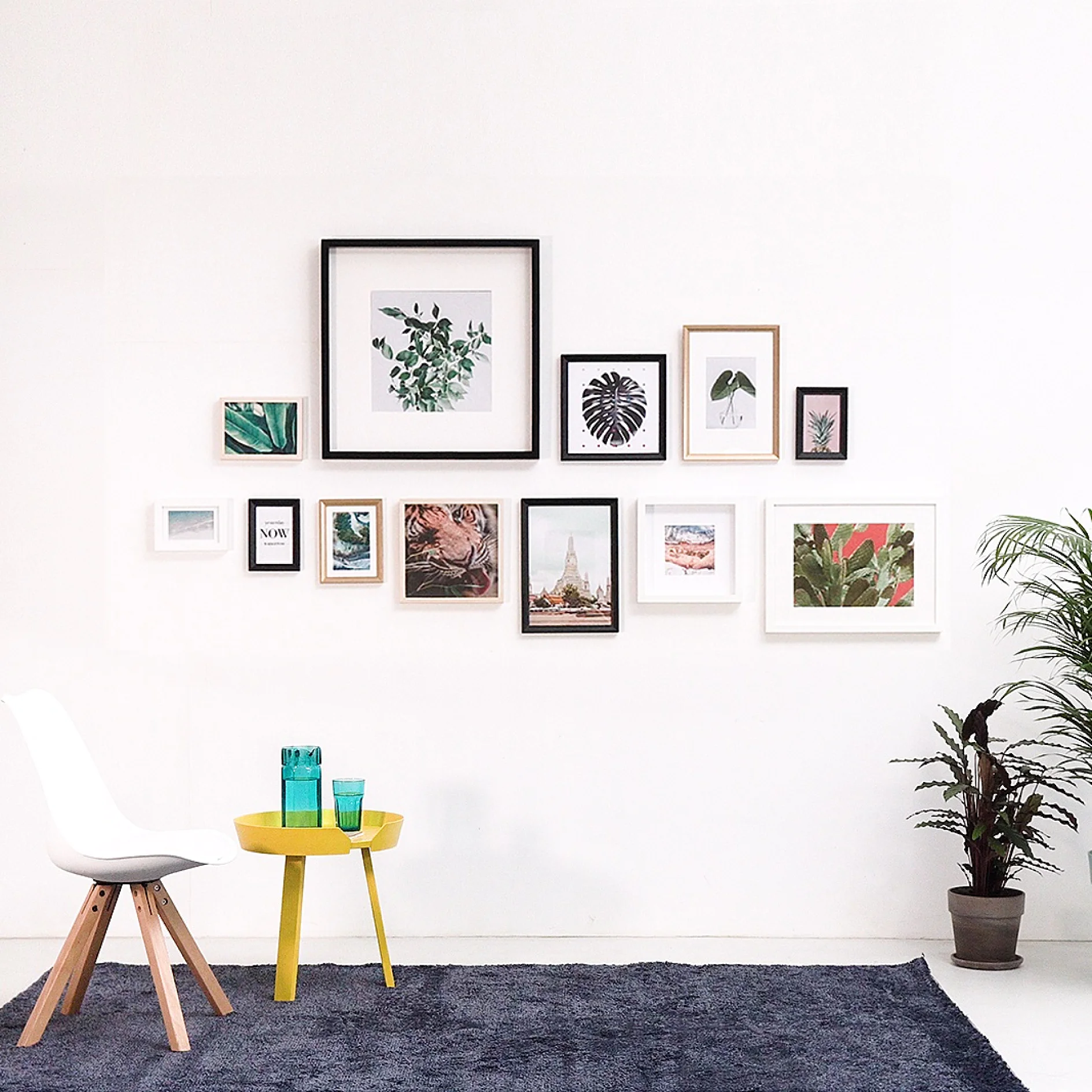 A minimalist room with a white wall displaying various framed botanical and abstract artworks. Below, a yellow side table with two blue glasses and a white chair are on a blue rug. A potted plant sits to the right, secured neatly using tesa tape for an organized look. (This text has been generated by AI)