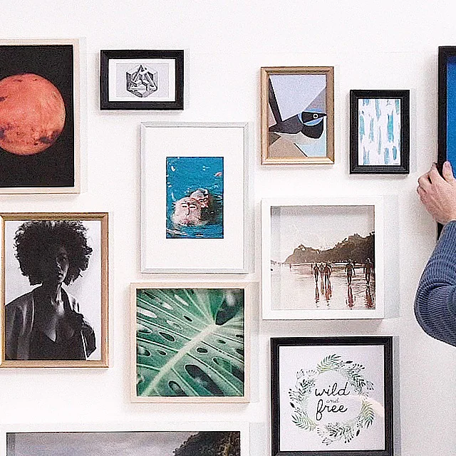 A person in a blue sweater is adjusting a framed picture of a large rose on a blue background on a gallery wall using tesa tape. The wall features various framed artworks, including a red planet, landscapes, abstract designs, and nature images. (This text has been generated by AI)