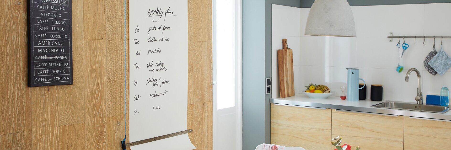 A modern kitchen with light wood cabinetry and a small dining table. A large notepad on the wall lists coffee drinks, held up securely by tesa tape. The table has a blue plate, bowl, and cup. Red flowers are in a vase, and fruits are on the counter. A pendant light hangs above. (This text has been generated by AI)