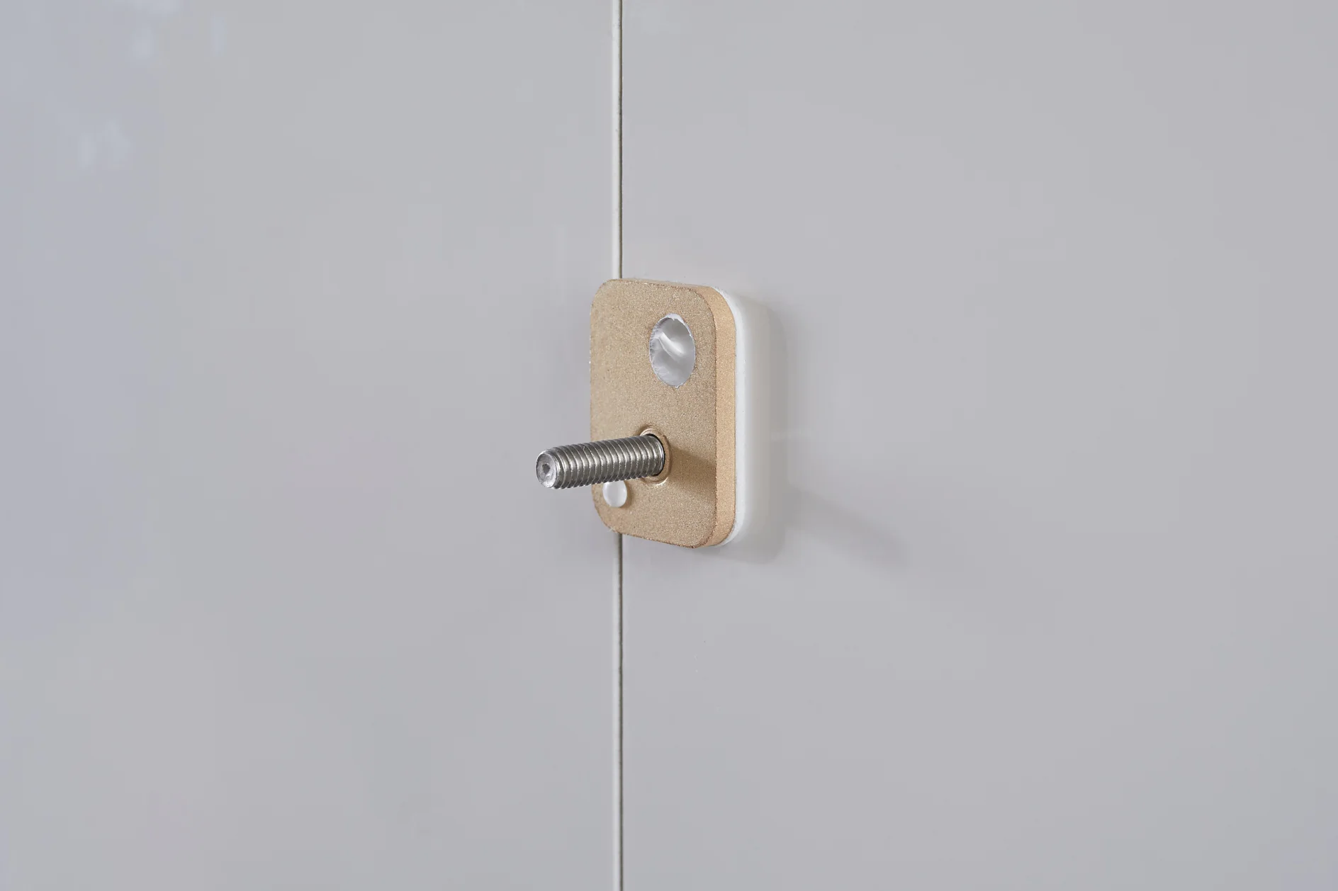 A close-up of a white surface with a rectangular wooden block attached using tesa tape. A metal threaded bolt protrudes from the block. The surface has a vertical seam, and theres a small, circular indentation above the block. (This text has been generated by AI)