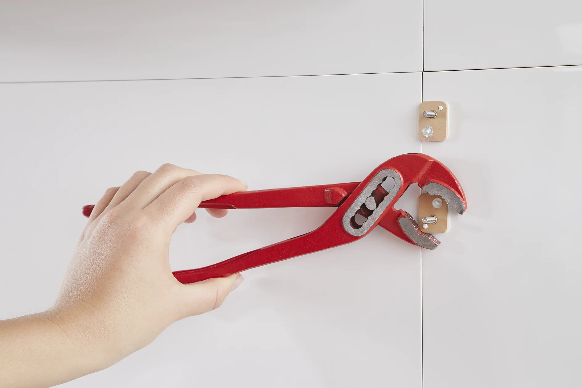 A hand uses red pliers to grasp a small gold clip attached to a white wall. The clip is held in place by screws and secured with tesa tape. (This text has been generated by AI)