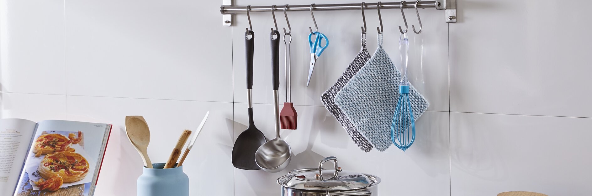 Modern kitchen with a stovetop and stainless steel cookware. Utensils hang from a rod on the wall, secured with tesa tape. An open cookbook and assorted vegetables are on the counter, fastened in place using tesa tape to prevent slipping. Frying pan, jars, and a blue vase with wooden utensils are also visible. (This text has been generated by AI)
