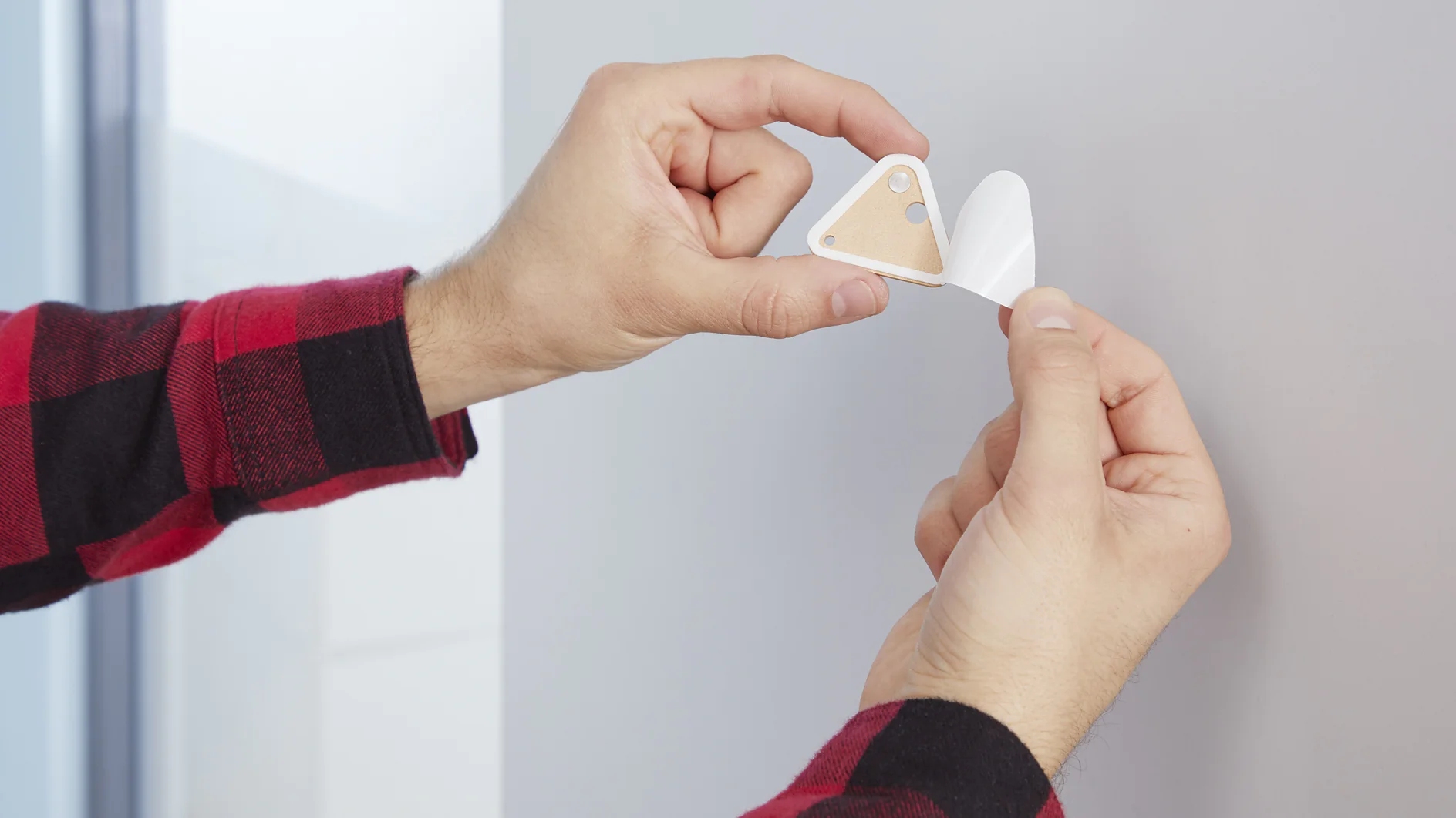 A person in a red and black plaid shirt is attaching a small triangular adhesive hook to a wall using tesa tape. The hook has a wooden accent and two holes. The hands are in focus, and the background is blurred. (This text has been generated by AI)