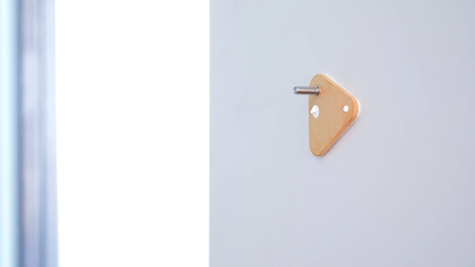 A close-up of a triangular yellow wall hook with a protruding metal pin, mounted on a light gray wall using tesa tape. The left side of the image shows a softly blurred vertical blue-gray edge. (This text has been generated by AI)