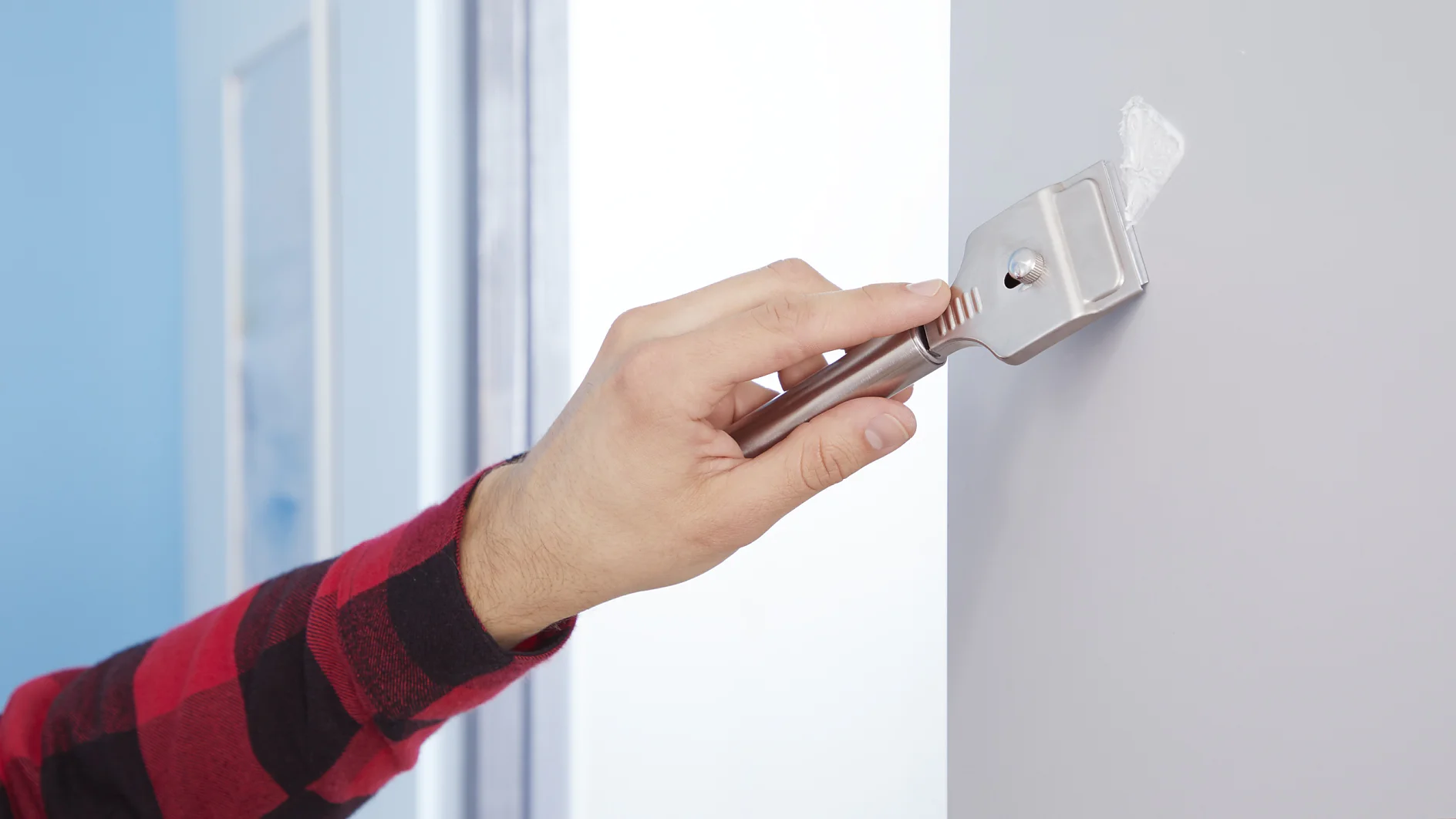 A person wearing a red and black plaid shirt is using a paint scraper to apply white filler to a hole in a gray wall. The scene is indoors, near a window, with natural light illuminating the area. (This text has been generated by AI)