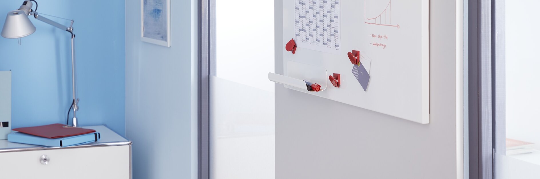 An office space with a light blue wall and a whiteboard mounted on a gray wall. The whiteboard features a monthly calendar and a small graph with red magnets held in place by tesa tape. A white chair and a lamp on a cabinet are visible. (This text has been generated by AI)