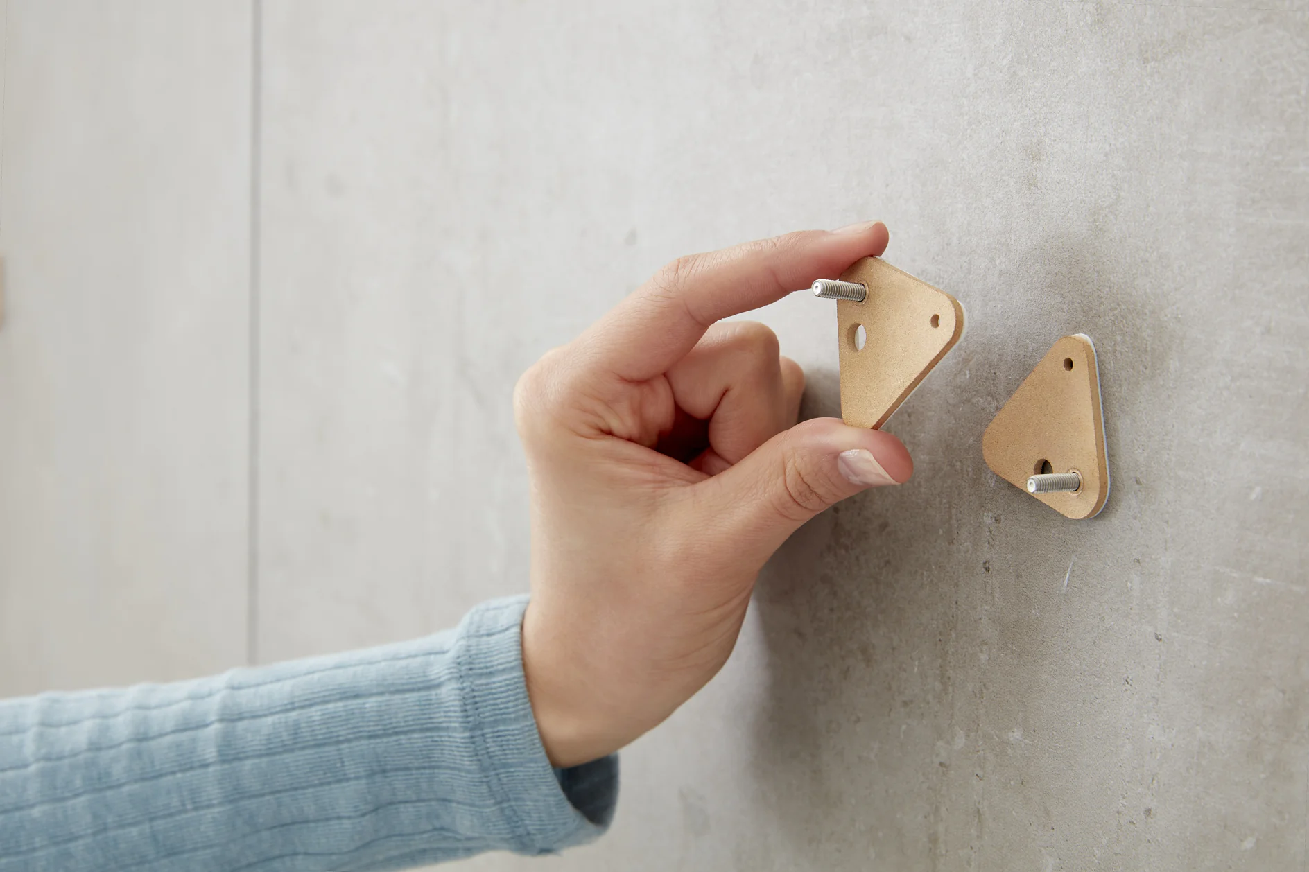 A hand in a blue sleeve attaches a triangular wooden object with a metal peg onto a gray wall using tesa tape. Another identical piece with a peg is already fixed on the wall. (This text has been generated by AI)