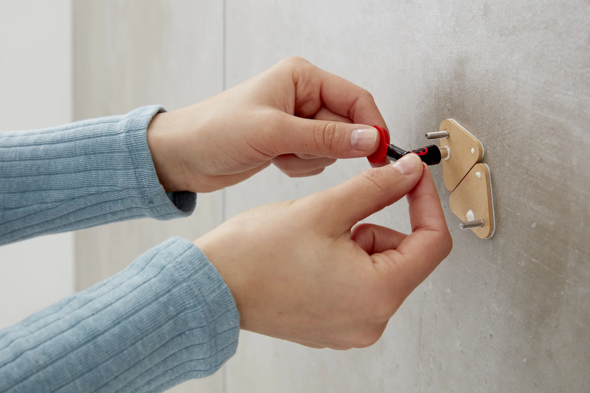A person in a light blue sweater is using a red-handled screwdriver to tighten a screw on a wall bracket with two hooks, secured with tesa tape. The wall has a smooth, light-colored surface. (This text has been generated by AI)