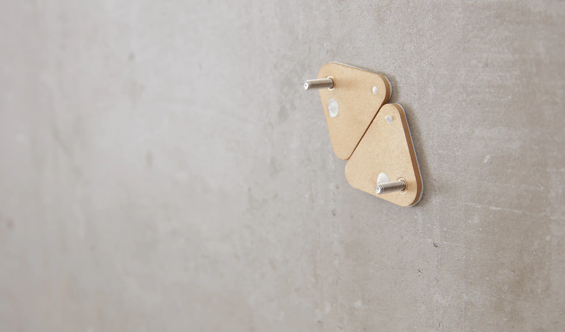 Two triangular cardboard pieces with metal pegs are mounted on a smooth, gray wall using tesa tape. The objects are positioned closely together near the center of the image. The background is plain and unadorned. (This text has been generated by AI)