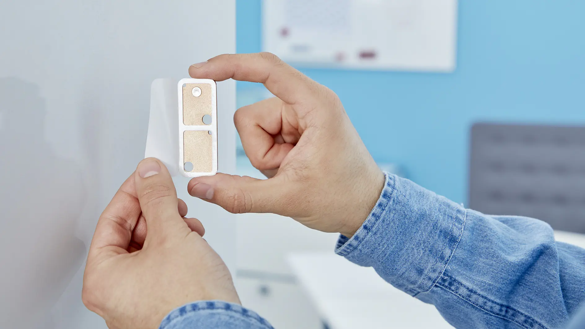 A person in a denim shirt is holding a white rectangular object with a tesa tape pad on one side and two magnets on the other. The background shows a blurred office setting with a whiteboard and a desk. (This text has been generated by AI)