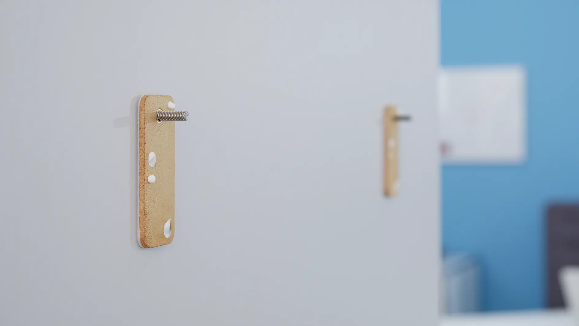 Two wooden wall brackets with protruding metal rods on a white surface, secured with tesa tape against a blurred background of a blue wall with a white item in the distance. (This text has been generated by AI)