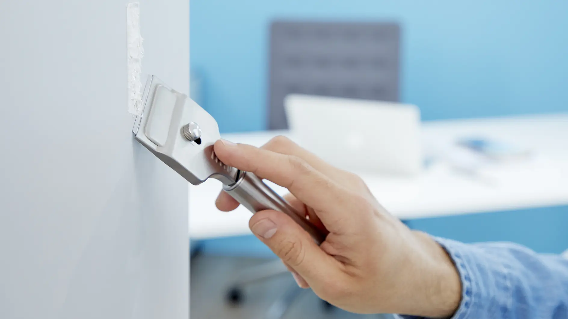 A person uses a metal scraper to remove material from a white wall. In the background, there is a blurred office setting with a chair and a laptop on a desk. The person is wearing a blue shirt, utilizing tesa tape for the task at hand. (This text has been generated by AI)