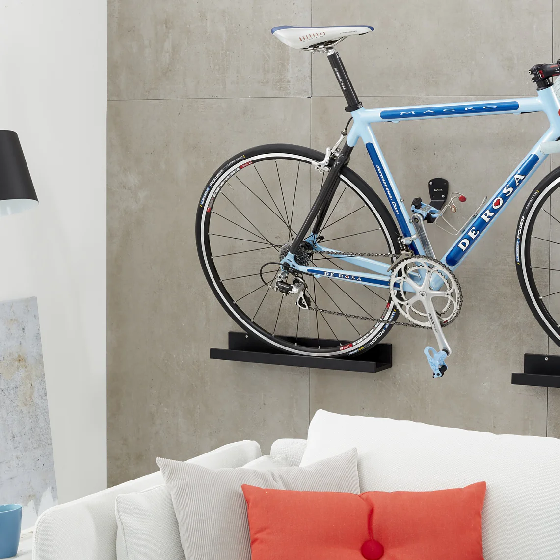 A blue and white road bicycle is mounted on a gray wall above a white sofa with colorful cushions, secured using tesa tape. A black floor lamp stands nearby, next to shelves with books and a painting. The setting appears to be a modern living room. (This text has been generated by AI)