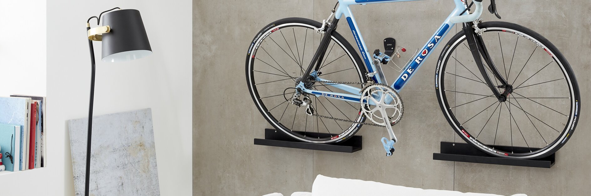 A blue and white road bicycle is mounted on a gray wall above a white sofa with colorful cushions, secured using tesa tape. A black floor lamp stands nearby, next to shelves with books and a painting. The setting appears to be a modern living room. (This text has been generated by AI)