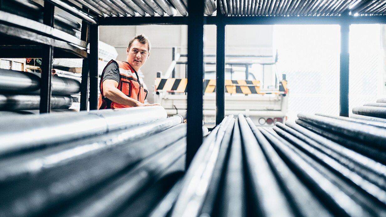 A person wearing an orange safety vest stands in a warehouse filled with metal pipes. They are looking to the side, surrounded by the stacked pipes on metal racks, with a truck partially visible in the background. (This text has been generated by AI)
