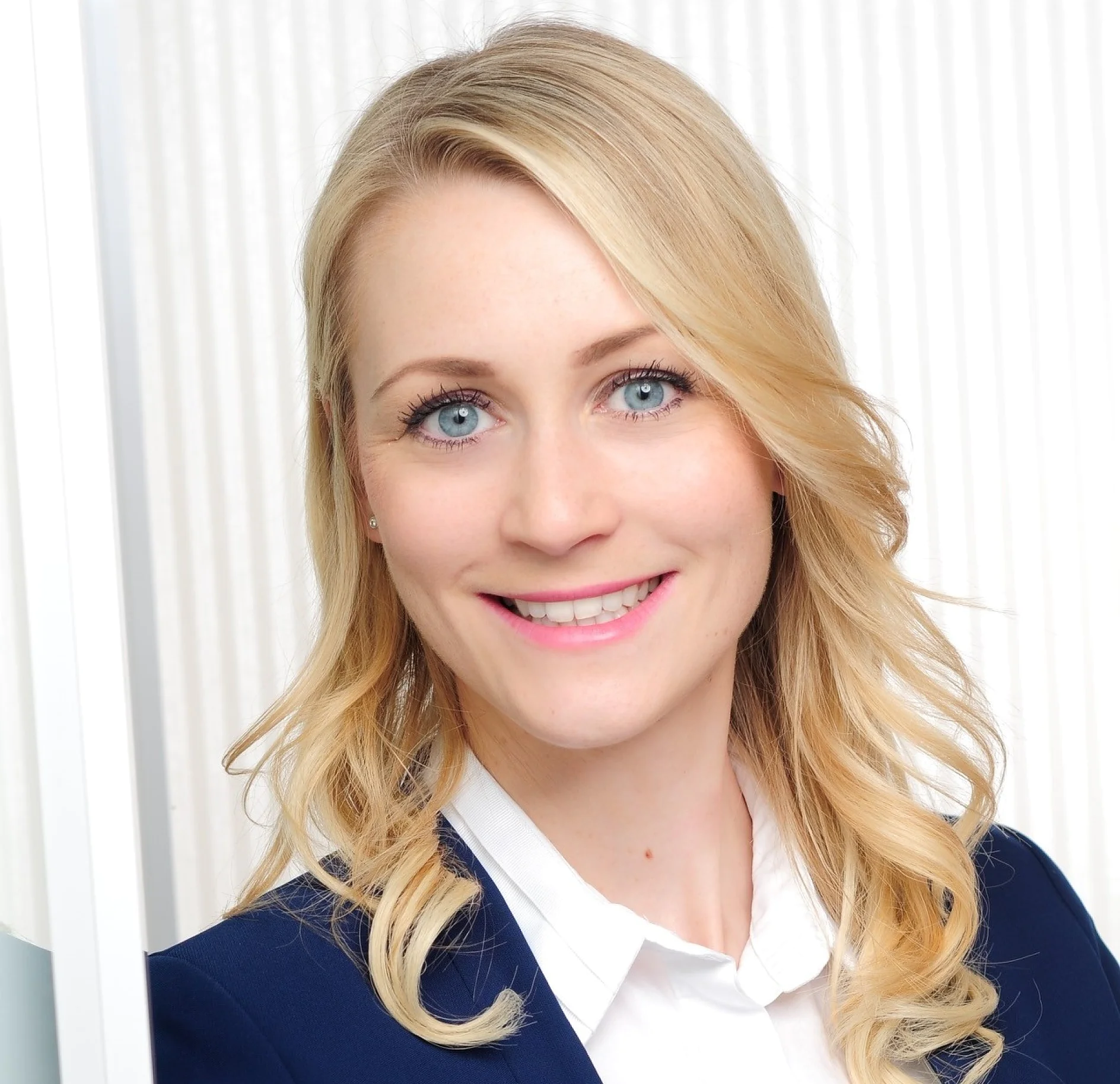 A person with long blonde hair is smiling at the camera. They are wearing a blue jacket over a white shirt. The background features a light-colored striped pattern. (This text has been generated by AI)