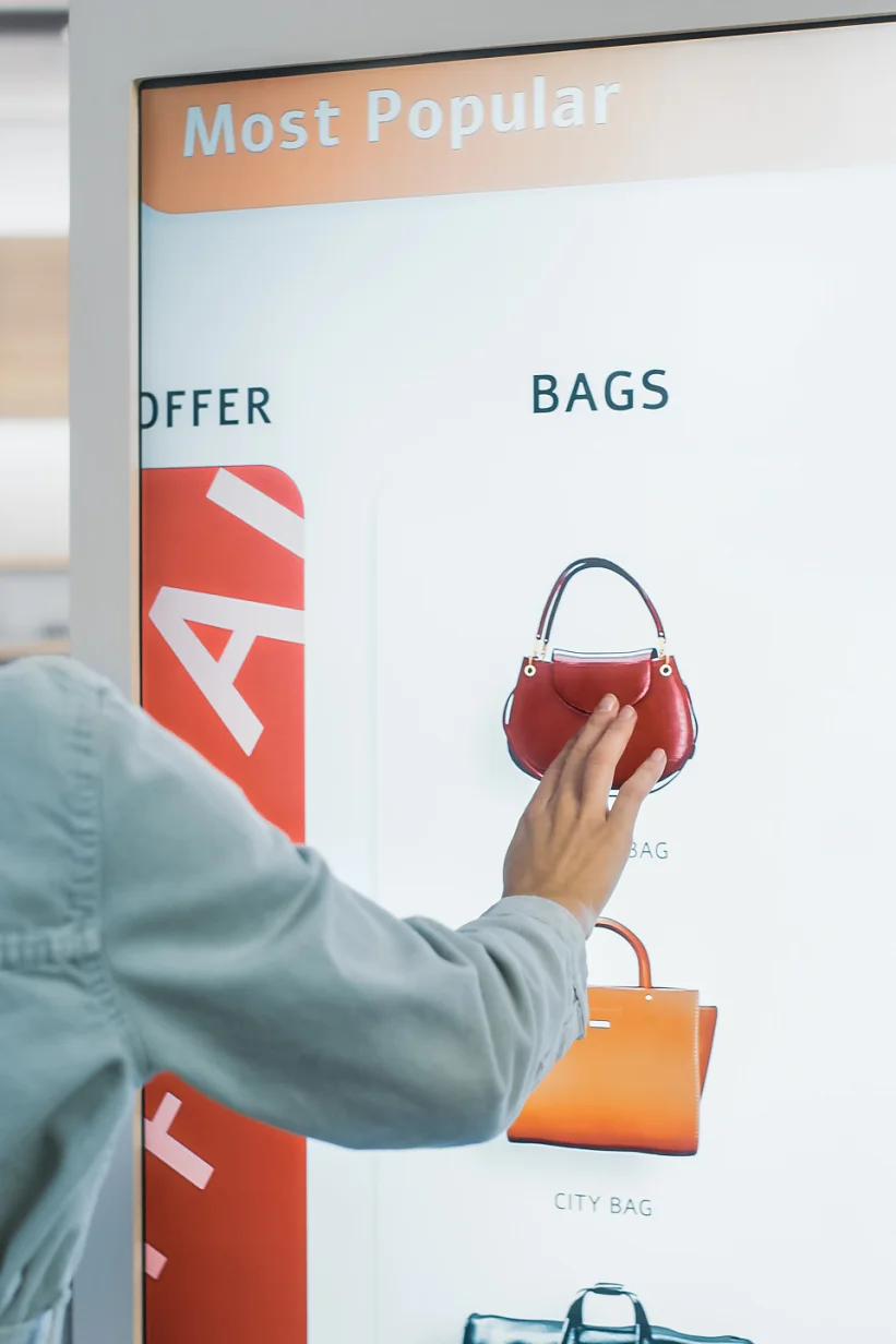 Cliente séduisante utilisant un écran tactile LCD au sol lors de ses achats dans un magasin de vêtements. Elle choisit des sacs élégants, sélectionne différents modèles de la collection. Personnes dans une boutique en vogue.