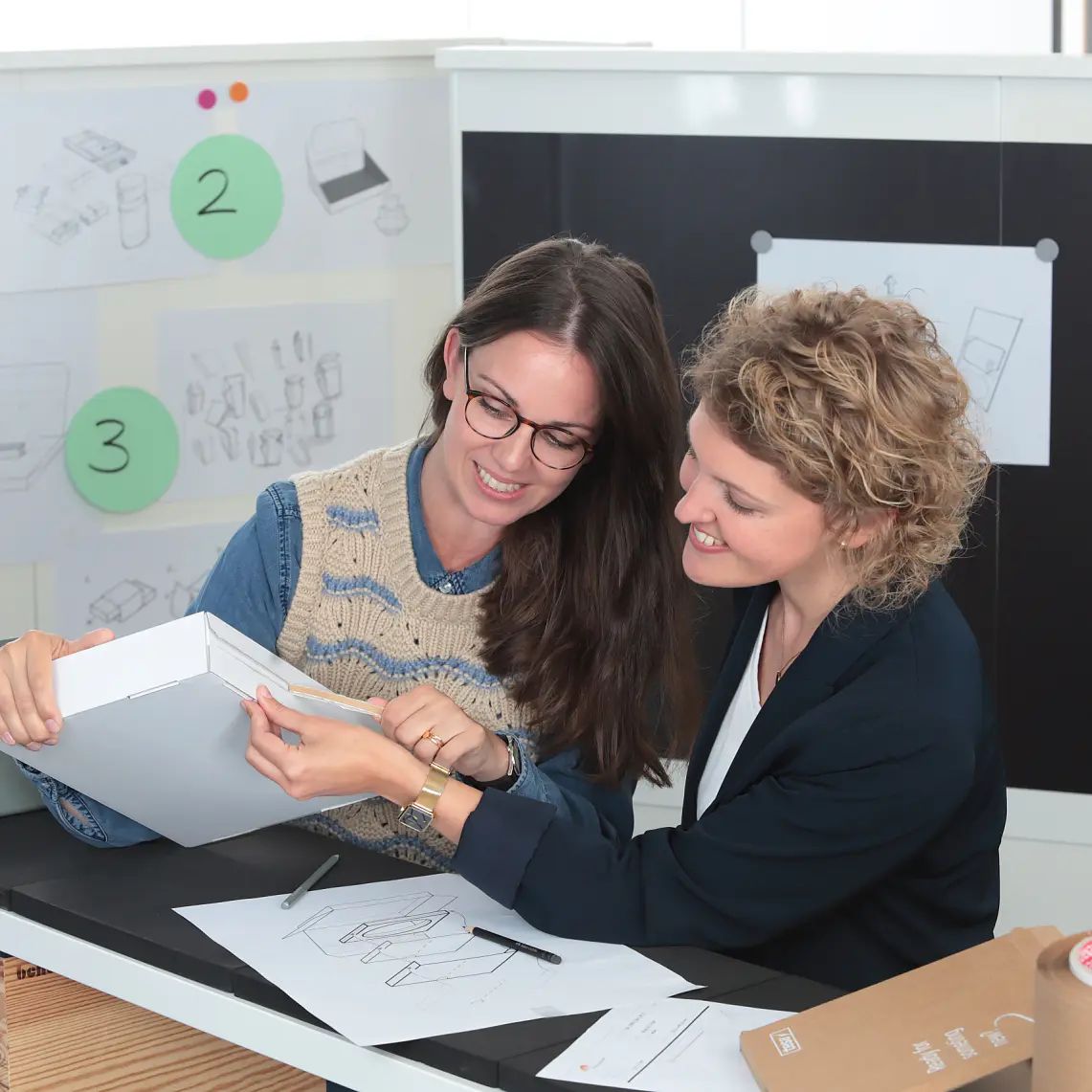 Two people are seated at a table with design sketches, examining a white box. The workspace has sketches and notes on the wall. Various office supplies, including tesa tape and markers, are on the table. Both appear engaged in discussion. (This text has been generated by AI)