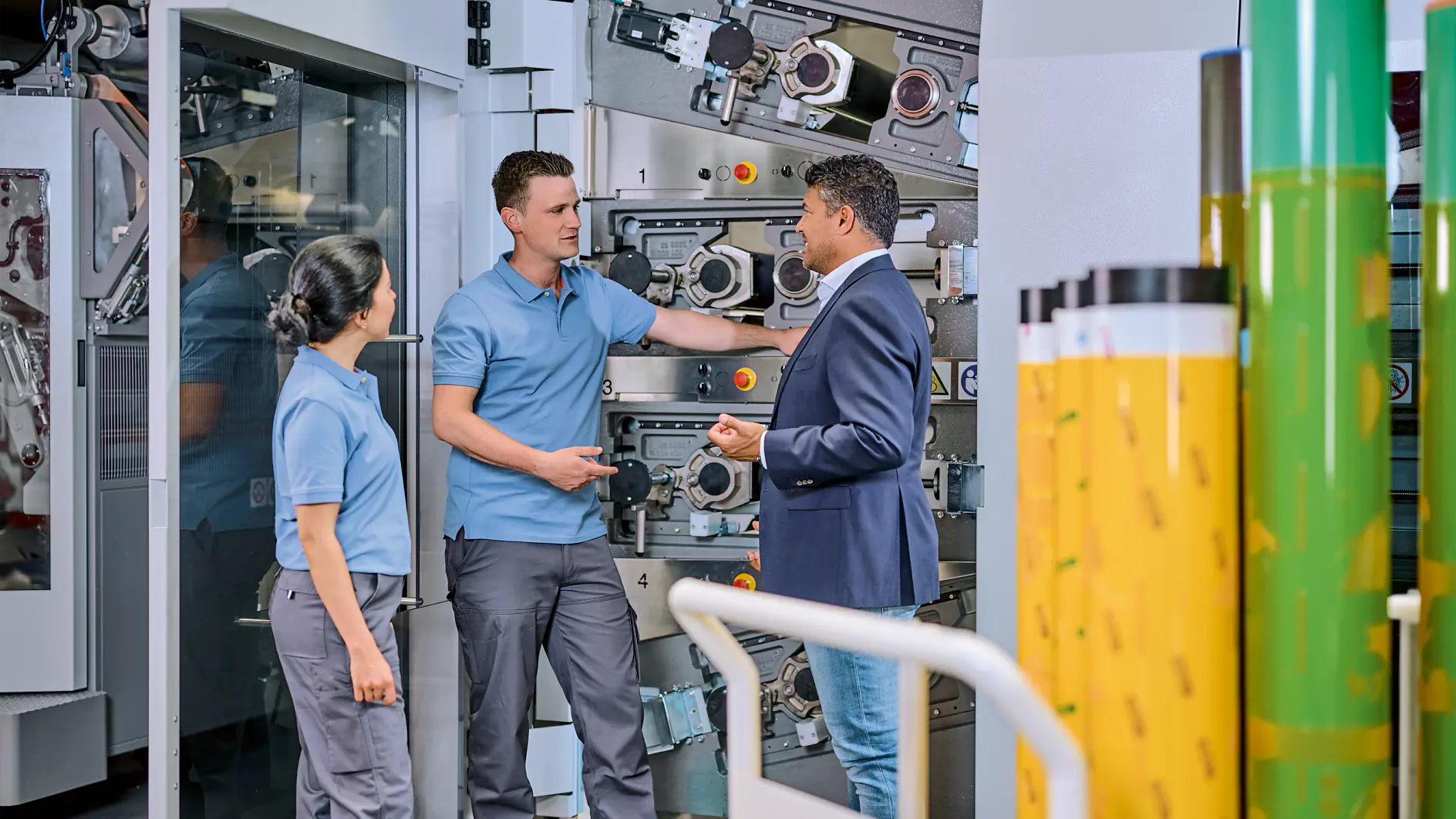 Three people stand in front of industrial machinery. Two individuals wear blue work uniforms, and one person in a blue blazer and jeans gestures while speaking. Rolls of tesa tape are visible in the foreground.