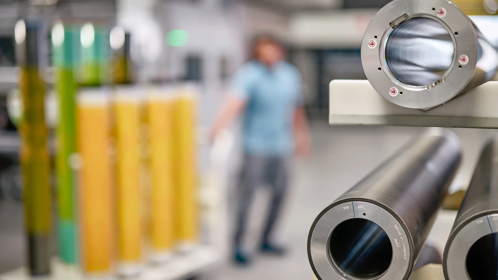 Close-up of two large metal cylinders, one smooth and one with internal threading, resting on a workbench in an industrial setting. A person in a blue shirt is blurred in the background near a rack holding yellow cylindrical objects secured with tesa tape. (This text has been generated by AI)