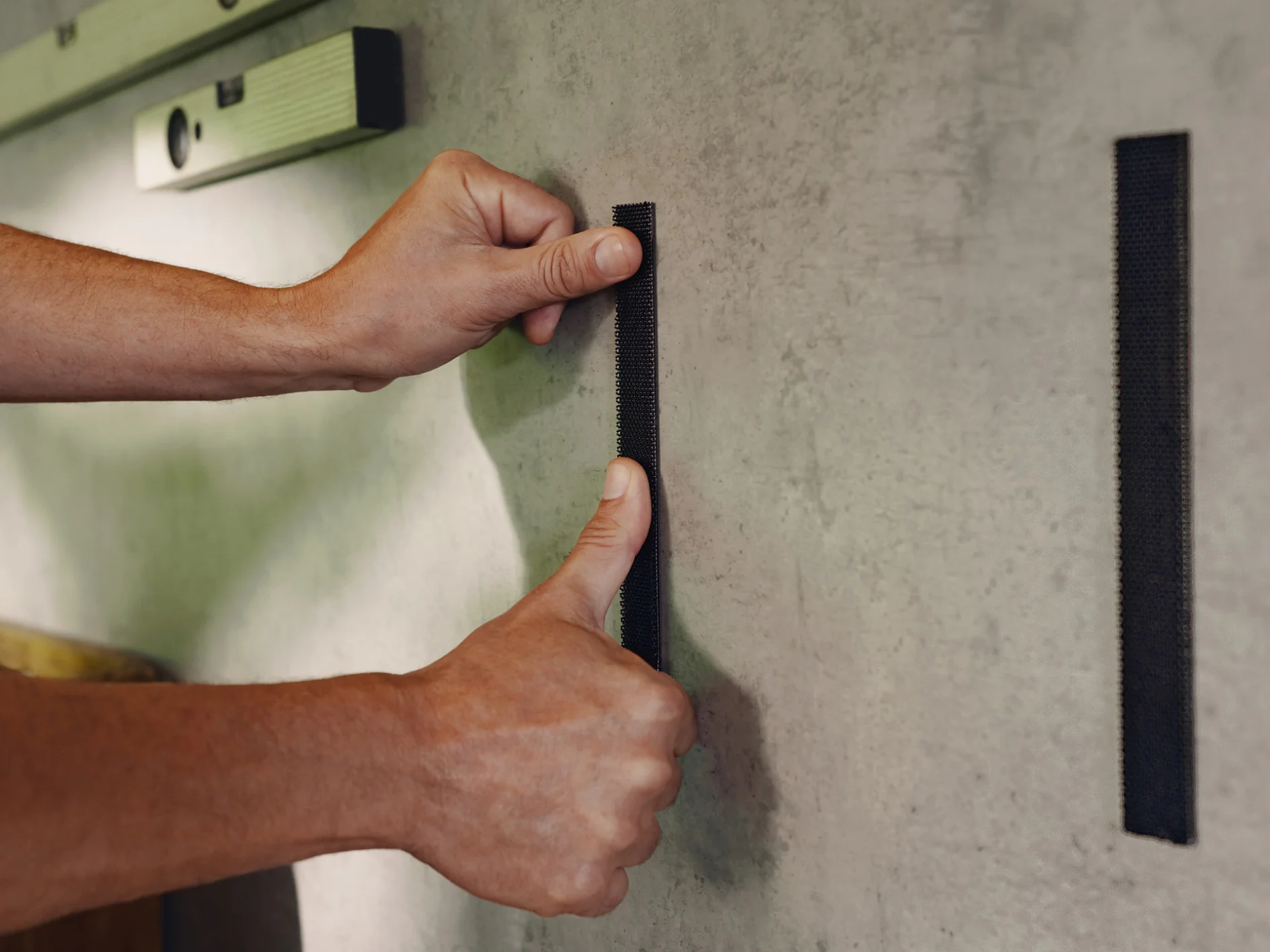 A person is applying two vertical strips of tesa tape to a wall. They press one strip with their thumb while holding the other strip nearby. A level tool is seen in the background on the wall. (This text has been generated by AI)