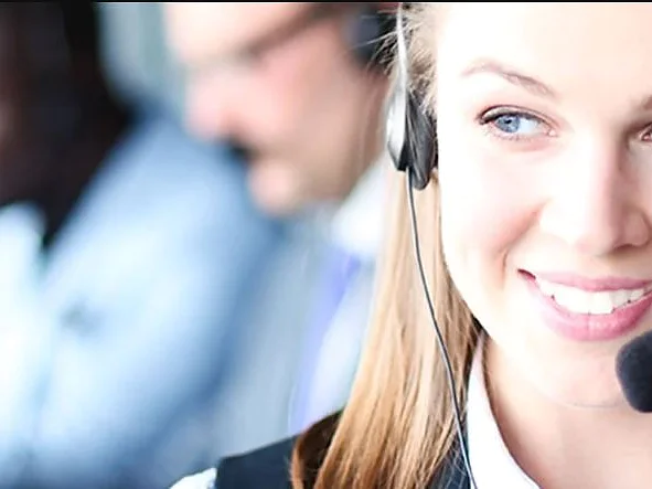 Une femme aux cheveux longs sourit et porte un casque avec microphone. Elle est nette au premier plan, tandis que deux autres personnes sont floues à larrière-plan, travaillant à un bureau avec des ordinateurs portables et utilisant du ruban adhésif tesa. (This text has been generated by AI)
