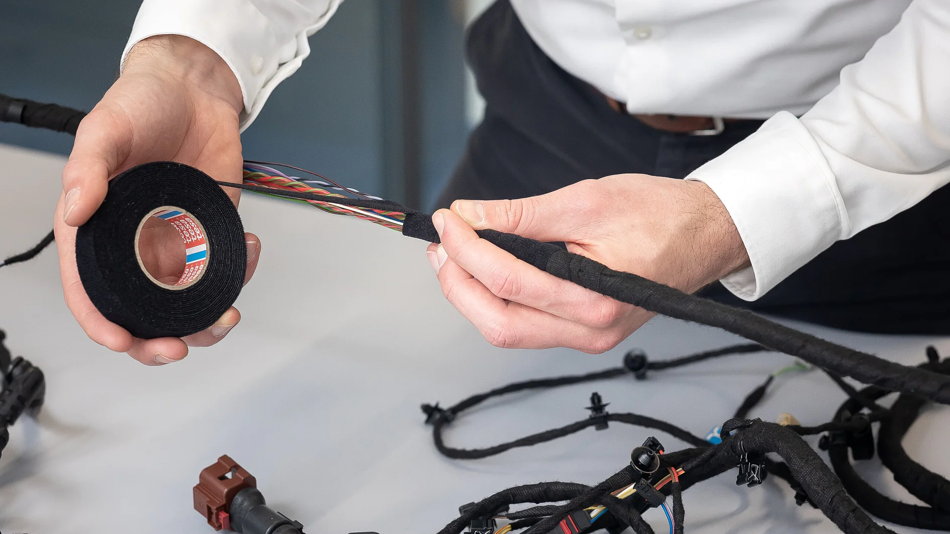 tesa tape being applied to an automotive wiring harness on a white table in an indoor workshop setting (This text has been generated by AI)
