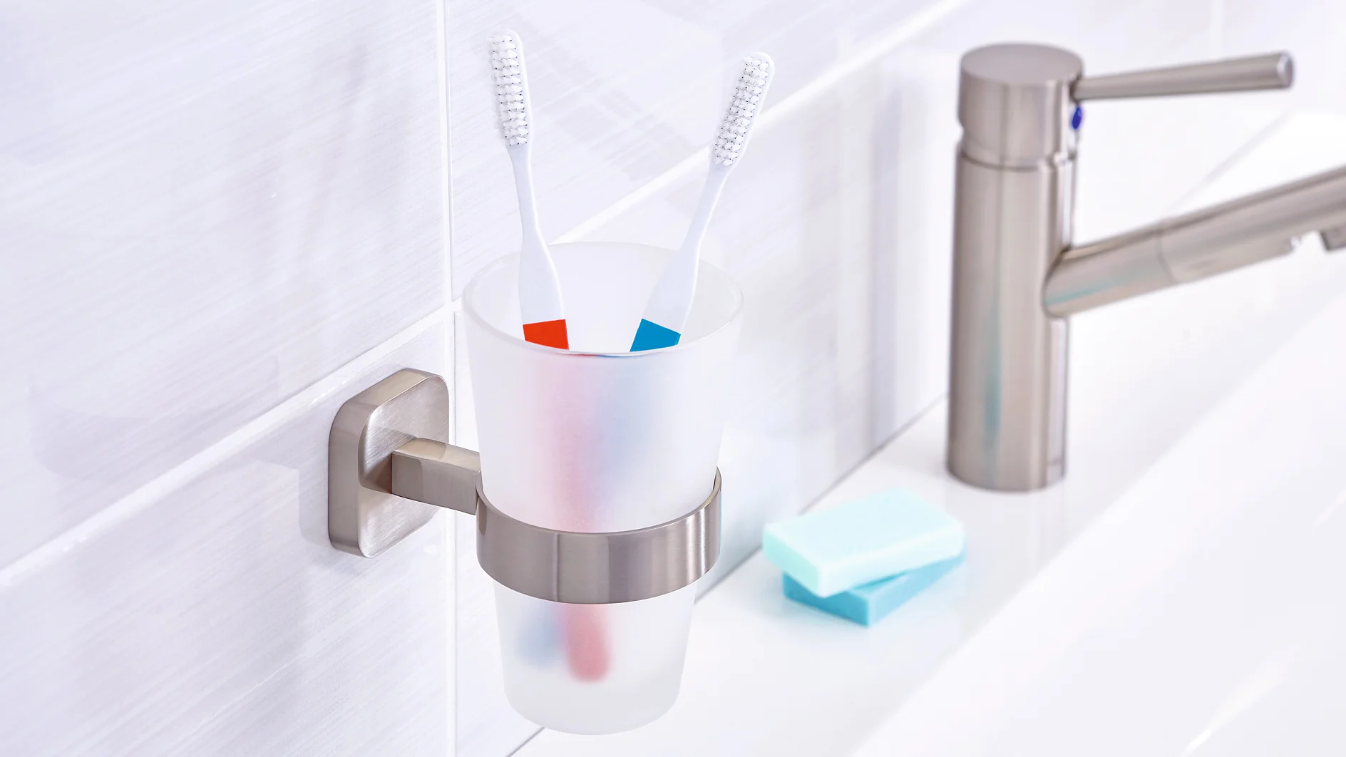 A bathroom scene featuring a matte white toothbrush holder attached to a wall with tesa tape, holding two toothbrushes with blue and red stripes. A silver faucet is on the right, along with a bar of soap on a white countertop. (This text has been generated by AI)
