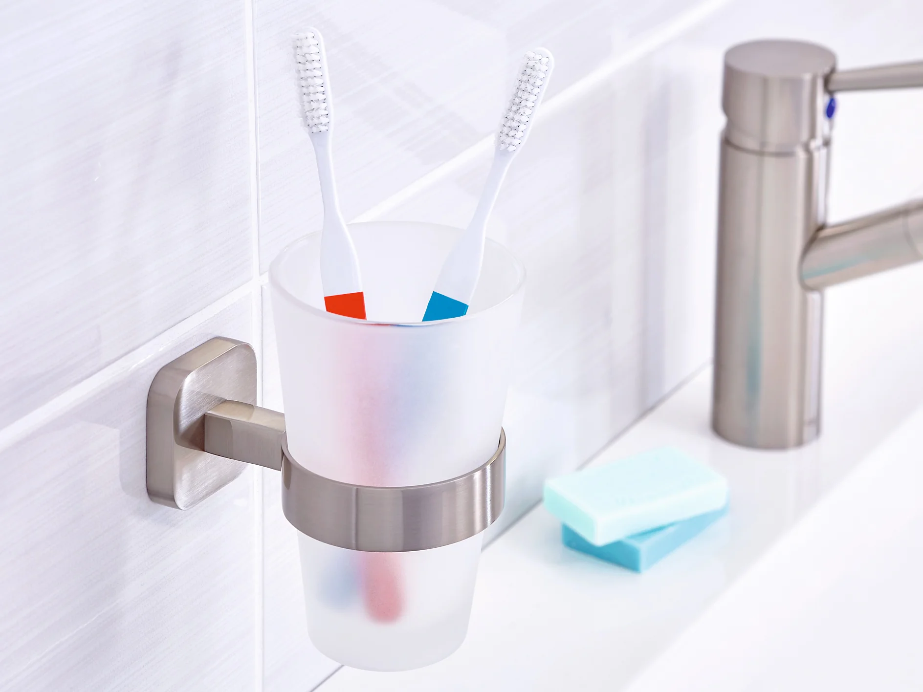 A bathroom scene featuring a matte white toothbrush holder attached to a wall with tesa tape, holding two toothbrushes with blue and red stripes. A silver faucet is on the right, along with a bar of soap on a white countertop. (This text has been generated by AI)