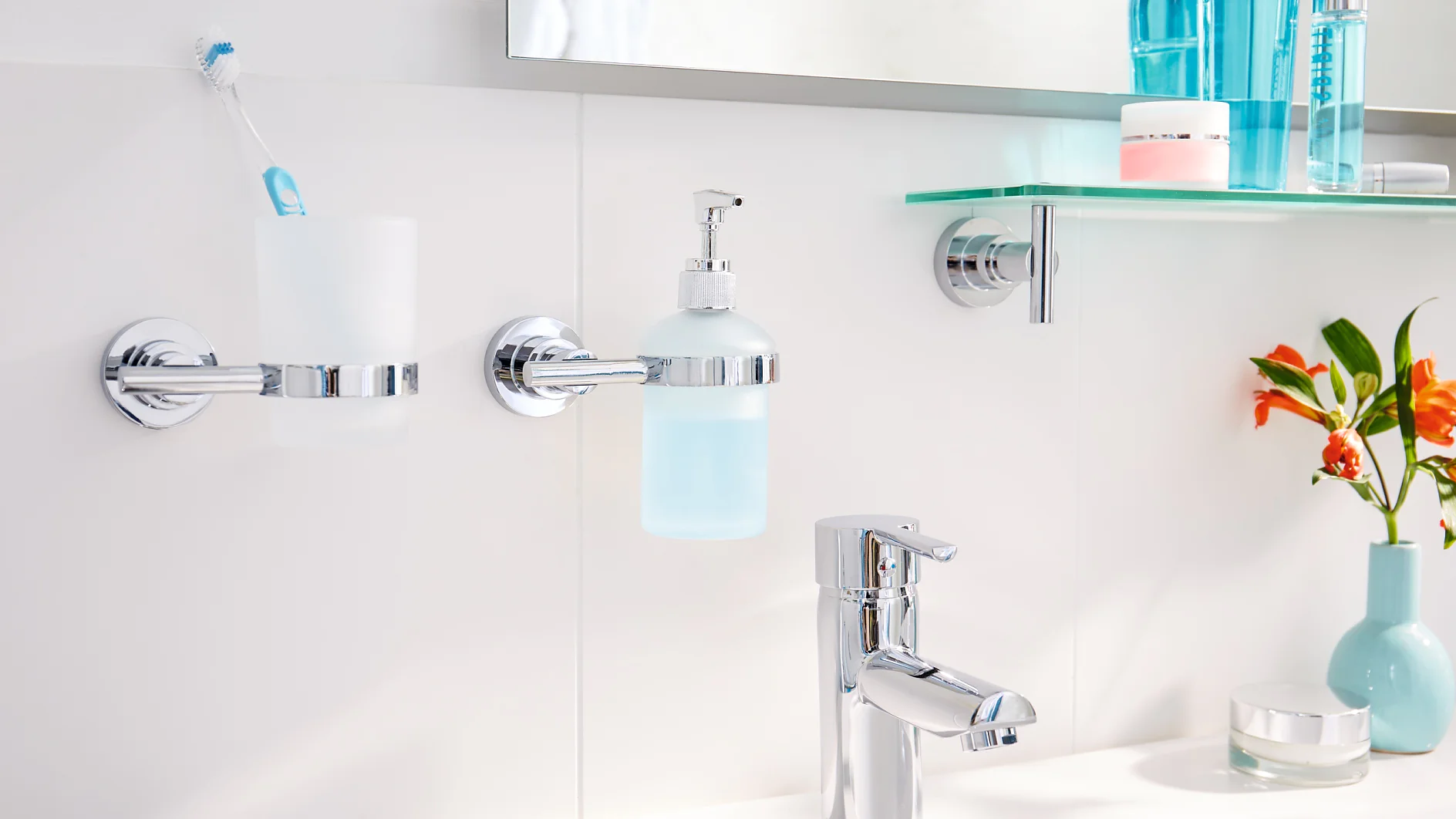 In the bathroom sink area, a chrome faucet complements a white counter. A toothbrush and holder, along with a soap dispenser, are securely mounted using tesa tape on the tiled wall. A shelf holding toiletries is also affixed with tesa tape. On the right side of the counter sits a small blue vase filled with orange flowers. (This text has been generated by AI)