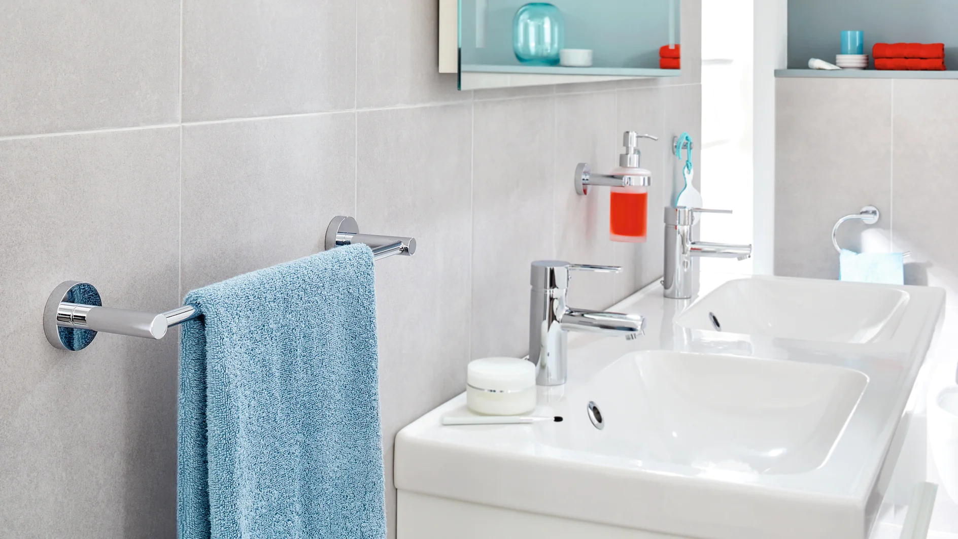 A modern bathroom features two white sinks with chrome faucets on a countertop. A blue towel hangs on a nearby towel bar, secured with tesa tape. A bright red soap dispenser is mounted on the wall using tesa tape. Shelves above are stocked with toiletries and towels, neatly held in place by tesa tape. (This text has been generated by AI)
