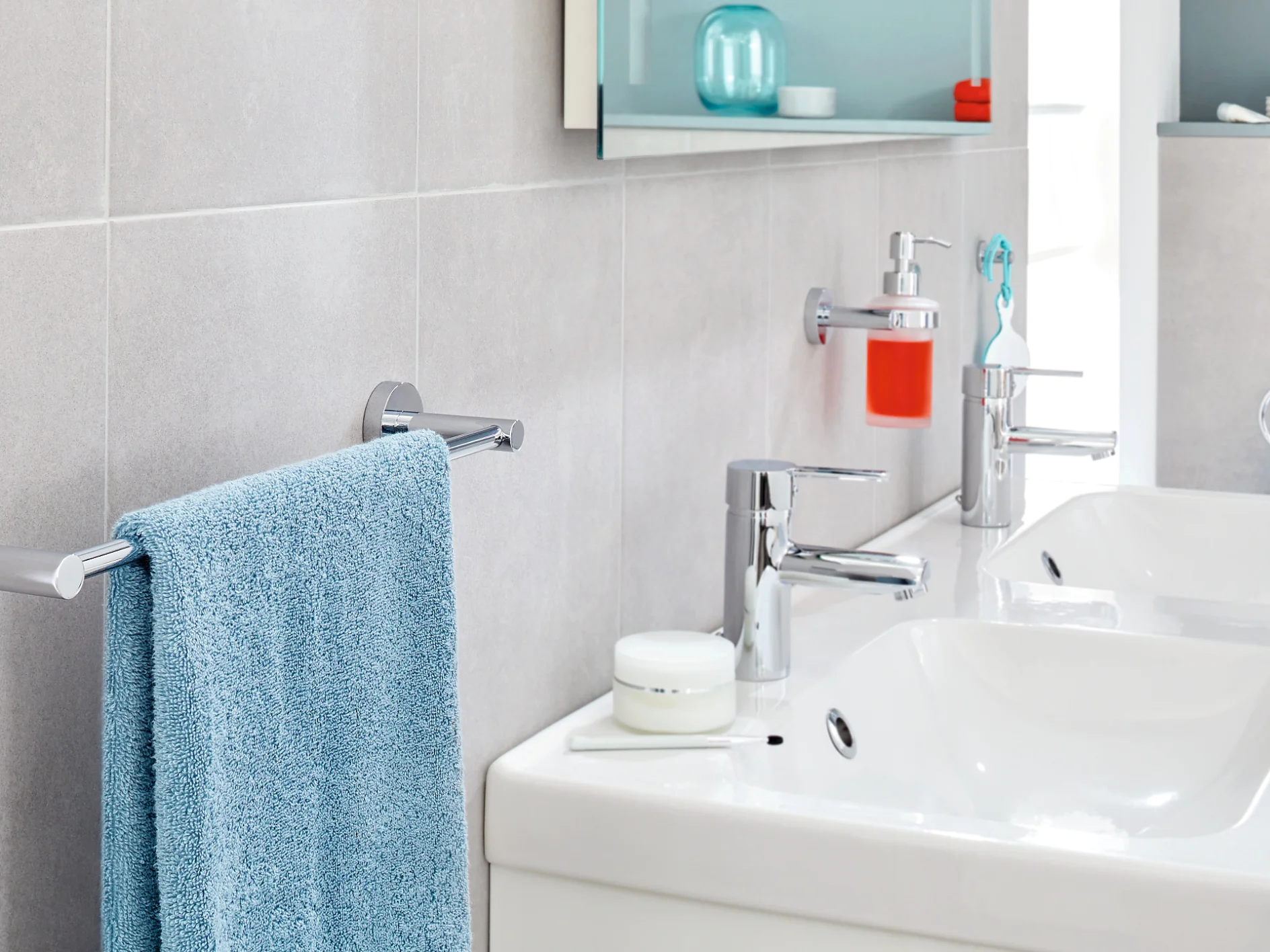 A modern bathroom features two white sinks with chrome faucets on a countertop. A blue towel hangs on a nearby towel bar, secured with tesa tape. A bright red soap dispenser is mounted on the wall using tesa tape. Shelves above are stocked with toiletries and towels, neatly held in place by tesa tape. (This text has been generated by AI)