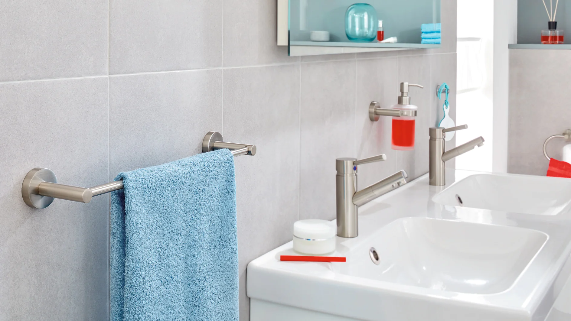 A modern bathroom sink area with two silver faucets, a white countertop, and a mounted soap dispenser containing red soap. A silver towel rack holds a blue towel, and shelves with toiletries are visible in the background. (This text has been generated by AI)