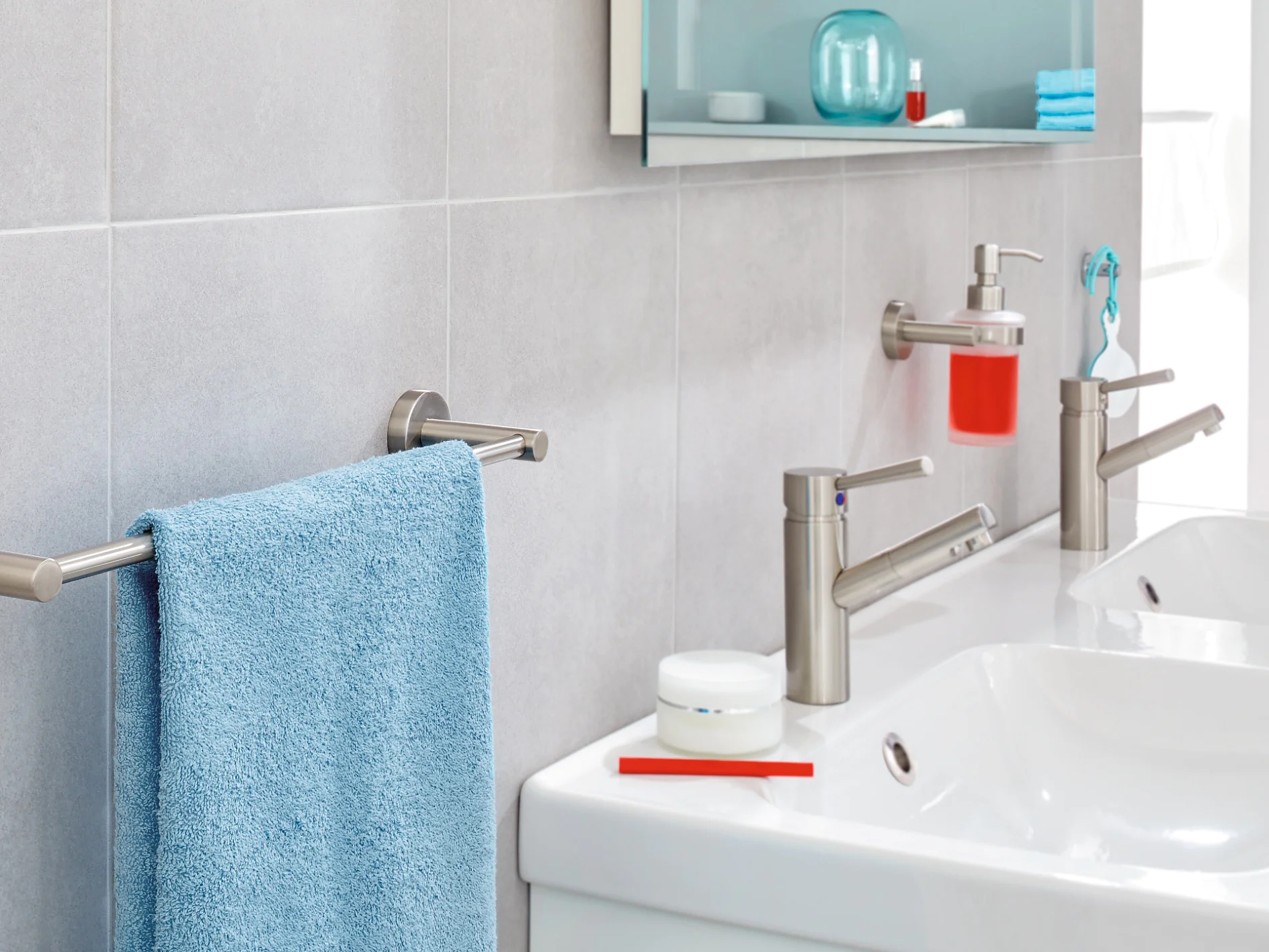 A modern bathroom sink area with two silver faucets, a white countertop, and a mounted soap dispenser containing red soap. A silver towel rack holds a blue towel, and shelves with toiletries are visible in the background. (This text has been generated by AI)