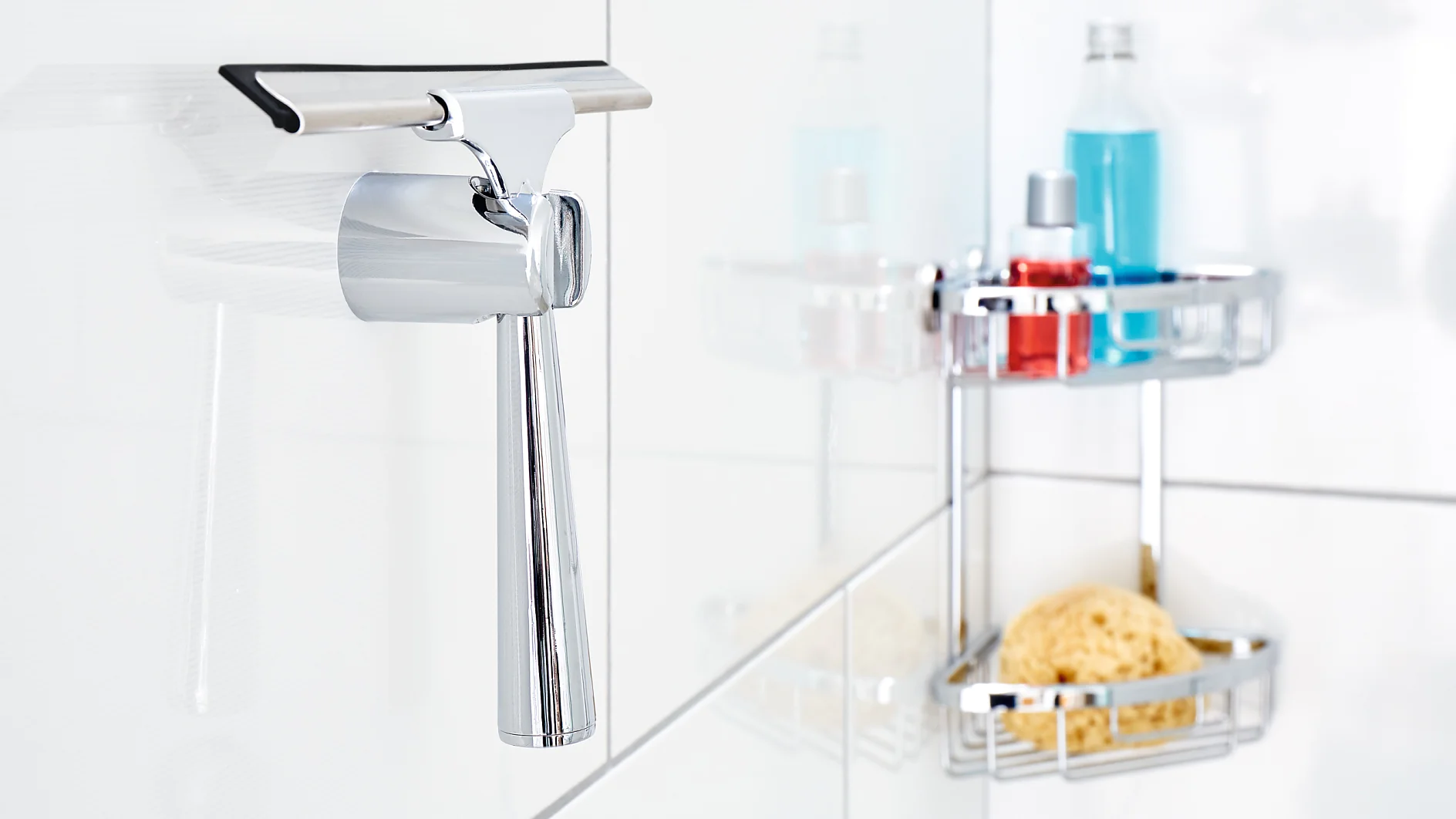 A chrome squeegee hangs on a white tiled bathroom wall, fastened securely with tesa tape. In the background, there are shower caddies holding a blue bottle, a red bottle, and a yellow sponge. (This text has been generated by AI)