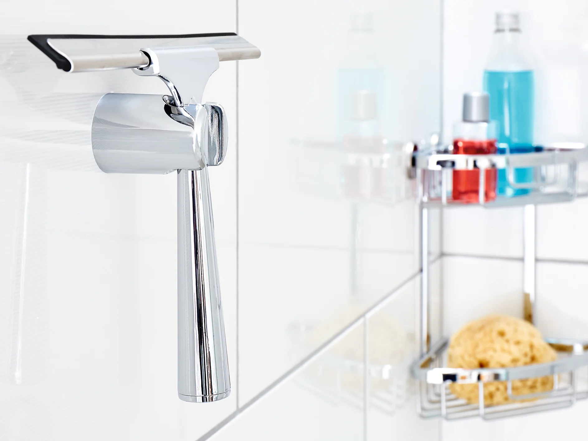 A chrome squeegee hangs on a white tiled bathroom wall, fastened securely with tesa tape. In the background, there are shower caddies holding a blue bottle, a red bottle, and a yellow sponge. (This text has been generated by AI)