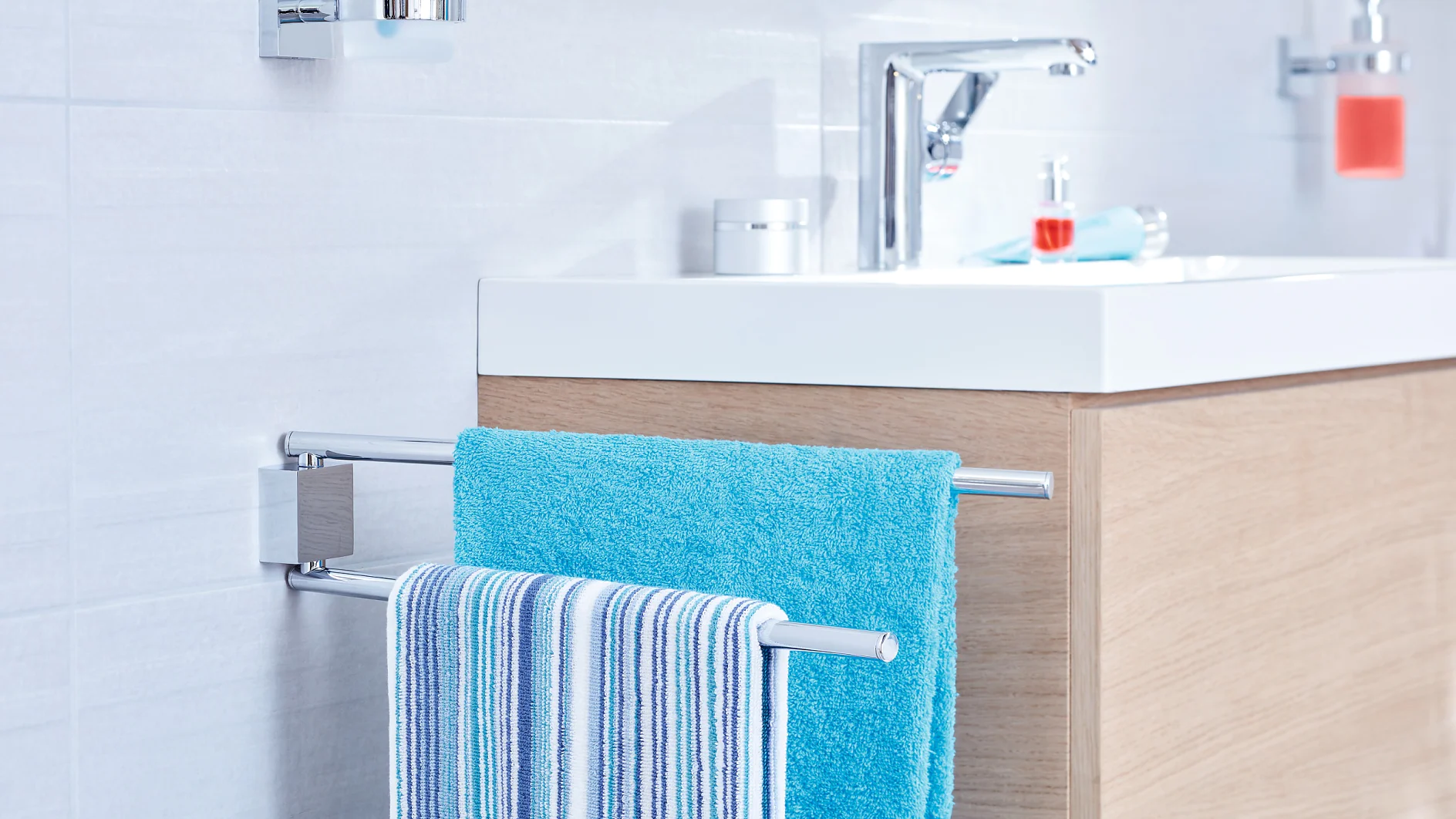 A modern bathroom features a white sink with a chrome faucet, set on a wooden cabinet. In the foreground, a towel rack holds a blue towel and a striped towel, mounted on the tile wall using tesa tape. (This text has been generated by AI)