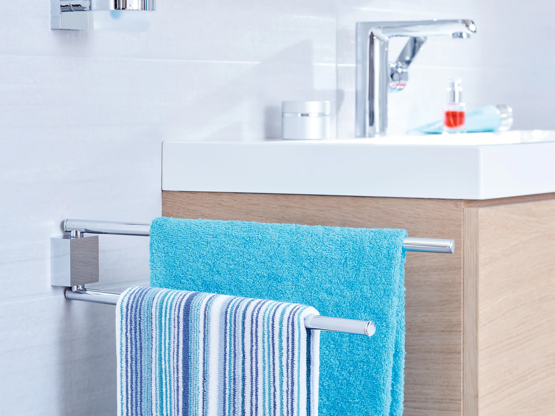 A modern bathroom features a white sink with a chrome faucet, set on a wooden cabinet. In the foreground, a towel rack holds a blue towel and a striped towel, mounted on the tile wall using tesa tape. (This text has been generated by AI)