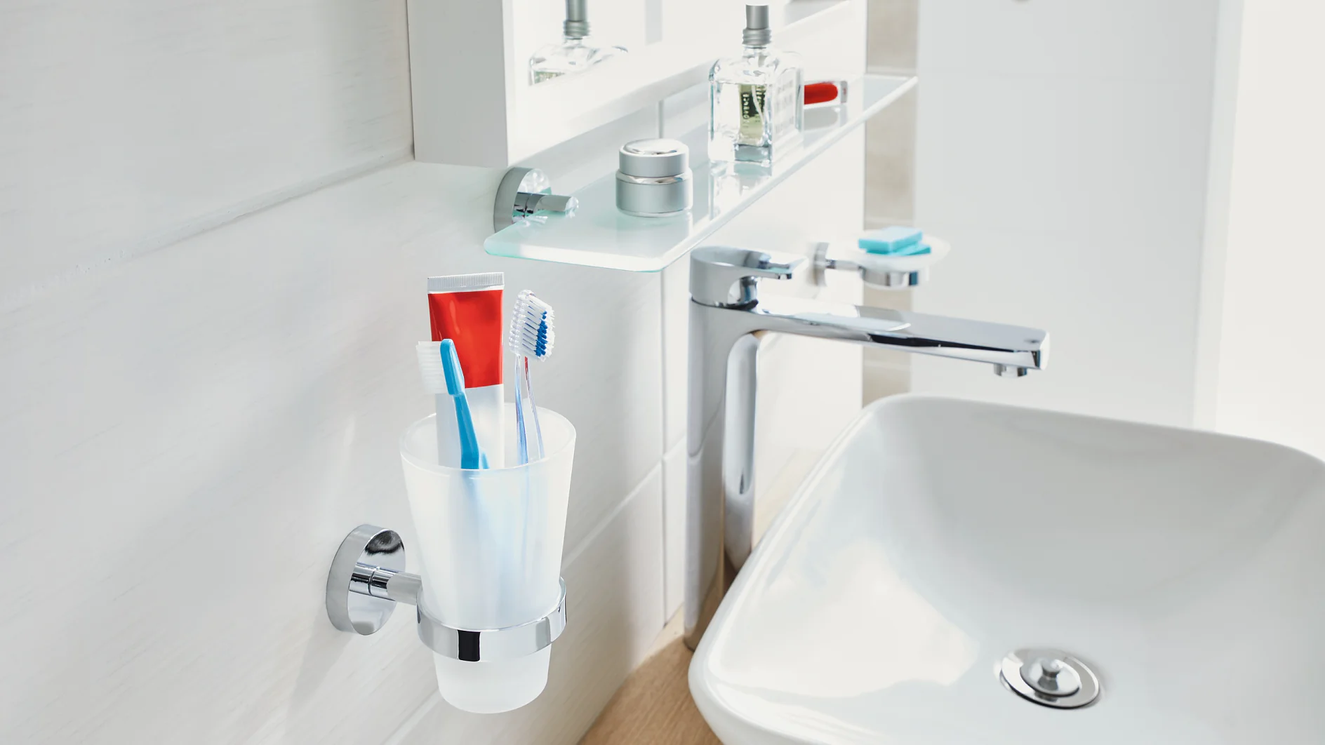 A bathroom sink area featuring a white basin with a silver faucet. A glass shelf above holds toiletries like cologne and creams. A wall-mounted cup contains two toothbrushes and a tube of toothpaste secured neatly in place using tesa tape. The background is white and clean. (This text has been generated by AI)