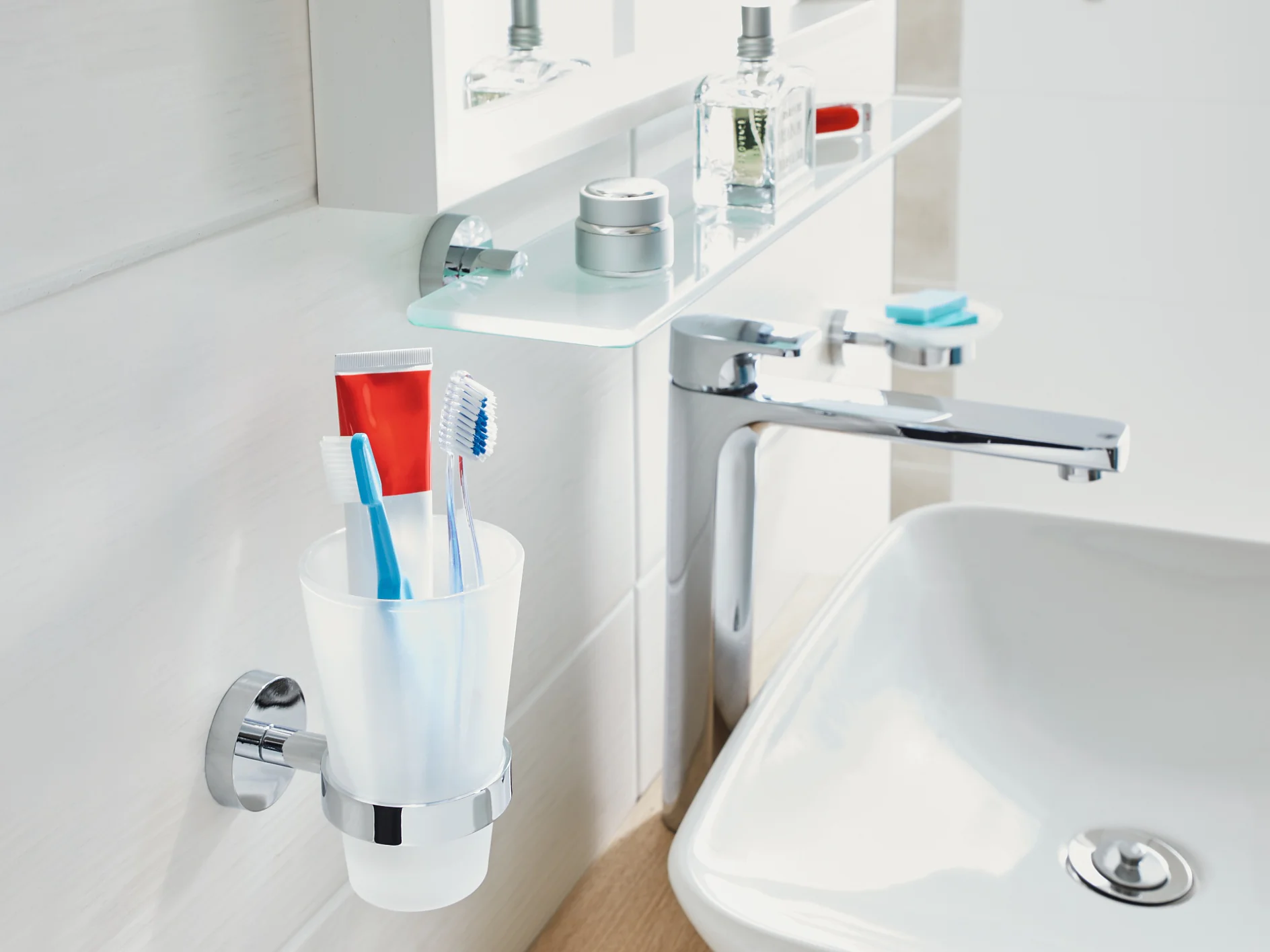 A bathroom sink area featuring a white basin with a silver faucet. A glass shelf above holds toiletries like cologne and creams. A wall-mounted cup contains two toothbrushes and a tube of toothpaste secured neatly in place using tesa tape. The background is white and clean. (This text has been generated by AI)