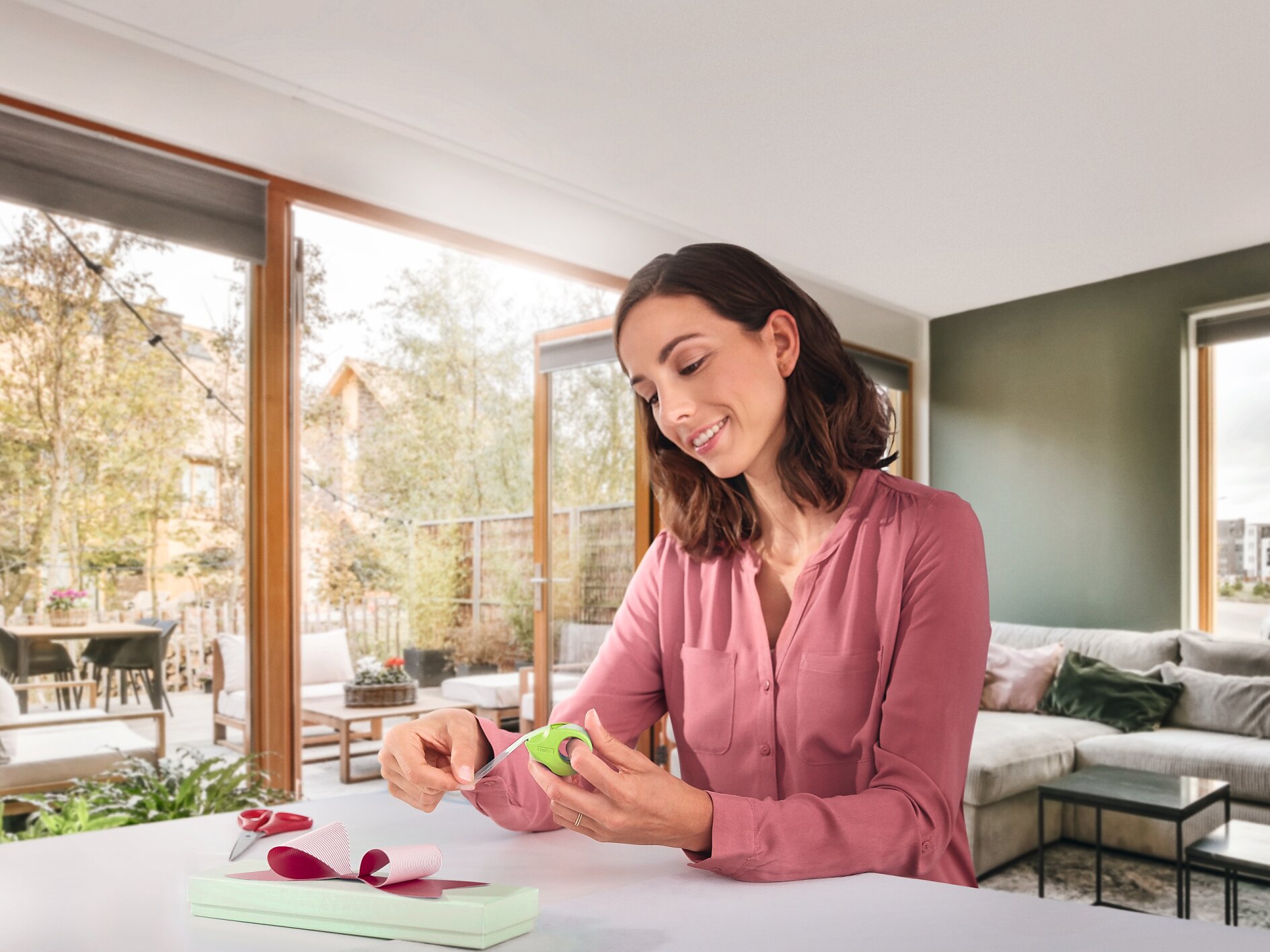 A woman in a pink blouse sits at a table in a modern living room, wrapping a box with green ribbon and tesa tape. Scissors and a spool of ribbon are on the table. Large windows and a patio are visible in the background. (This text has been generated by AI)