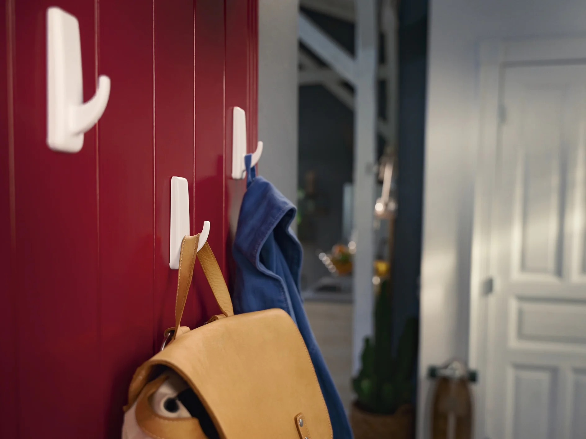 A red wooden wall with three white hooks. A tan leather bag hangs on the lower hook, secured by tesa tape, while a blue jacket hangs on the middle hook, also held up by tesa tape. A hallway and a white door are visible in the background. (This text has been generated by AI)